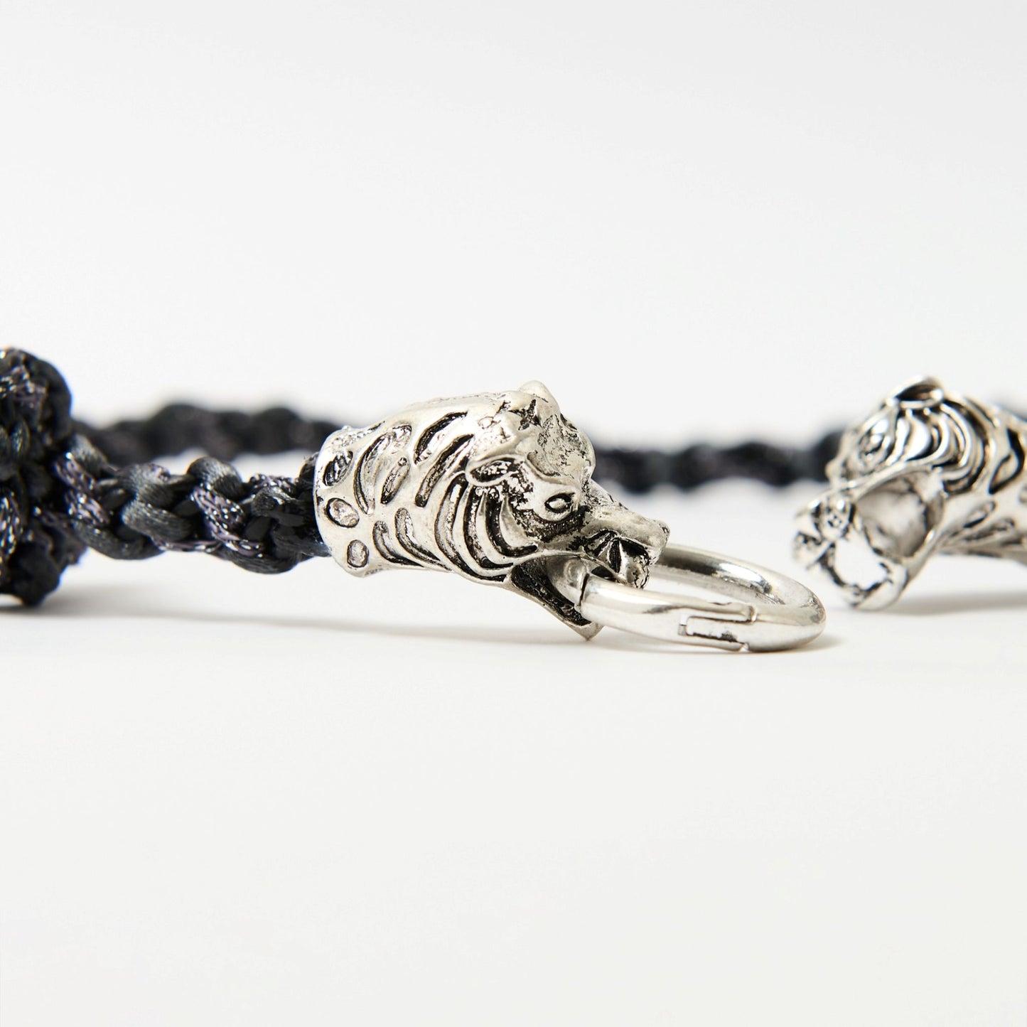 Close-up of a black braided cord necklace featuring a silver tiger head clasp with intricate detailing, each head gripping a central metal ring.