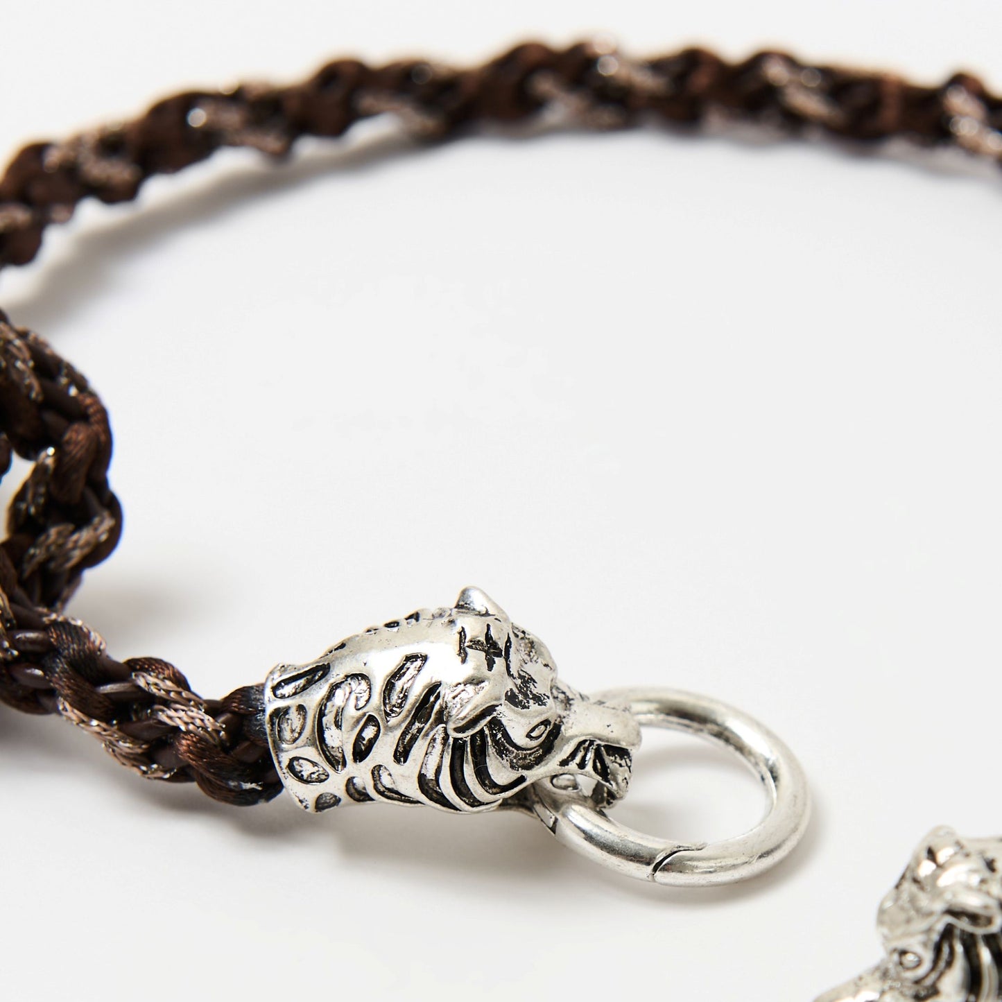 Close-up of a braided cord necklace in shades of brown featuring a silver tiger head clasp with intricate detailing, each head gripping a central metal ring.