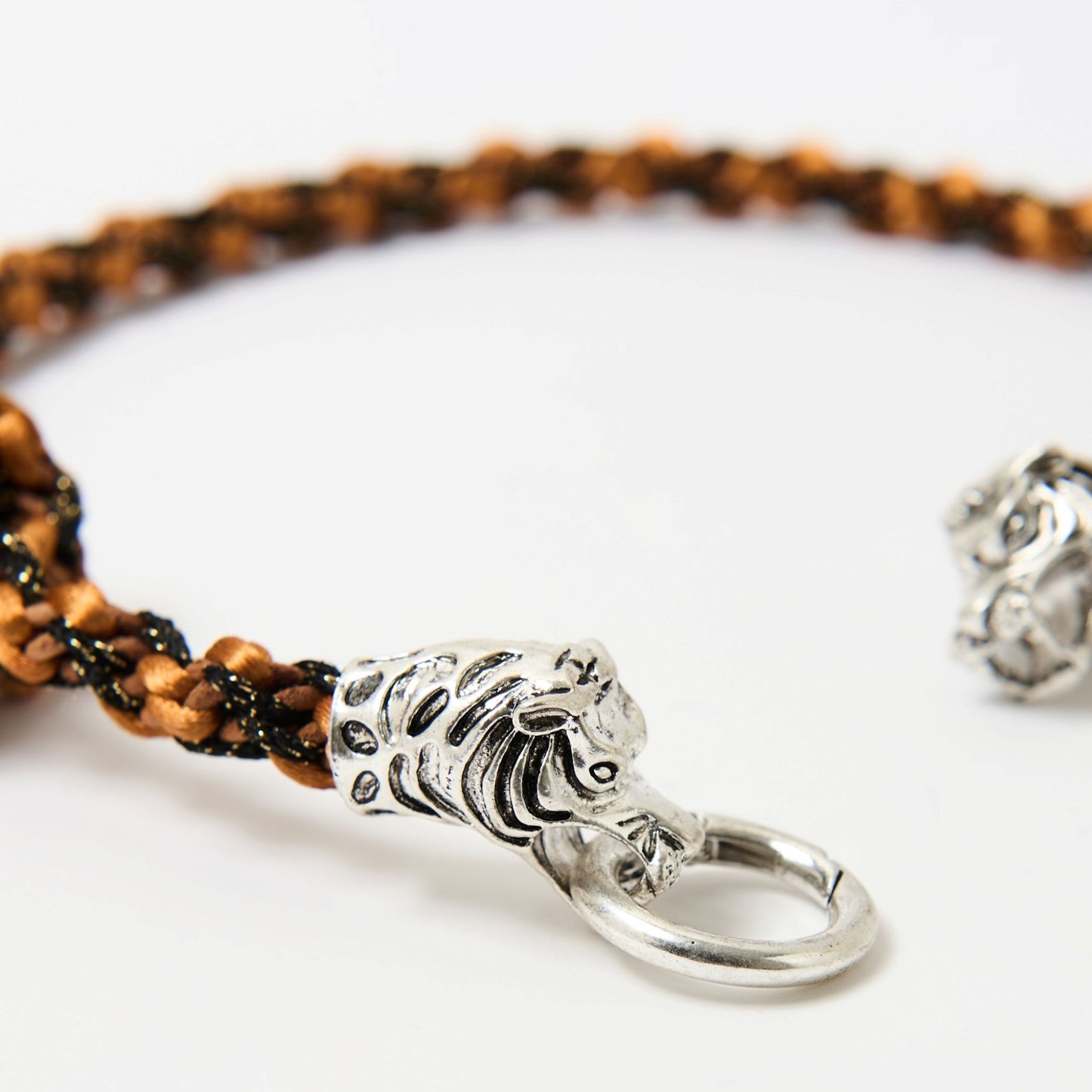 Close-up of a braided cord necklace in shades of orange and earthly brown featuring a silver tiger head clasp with intricate detailing, each head gripping a central metal ring.