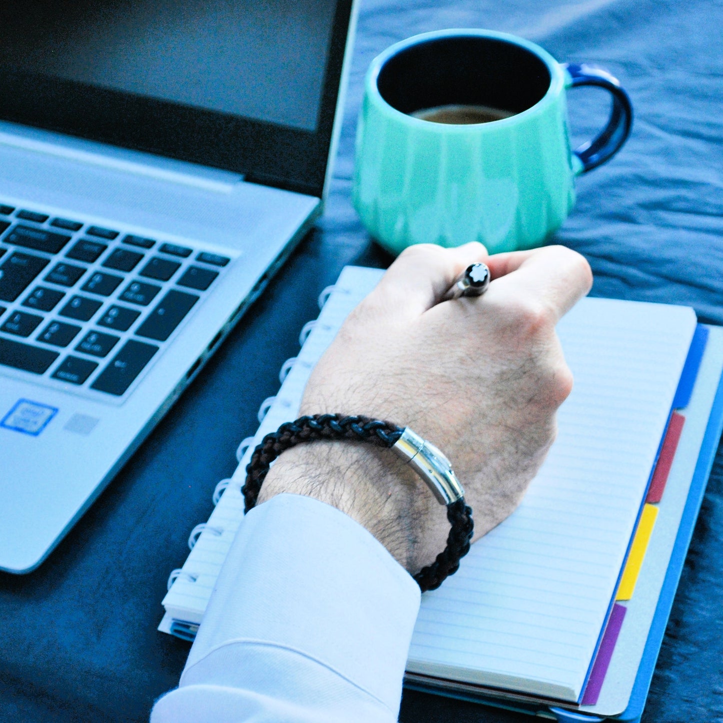 Person writing in a notebook with a laptop and mug on a table