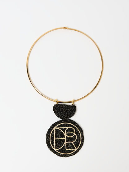 A minimalist gold choker necklace featuring a bold black textured pendant. The pendant has a circular design with an intricate gold geometric logo or pattern in the center, creating a striking contrast against the black background. The design is elegant and modern, ideal for statement styling.