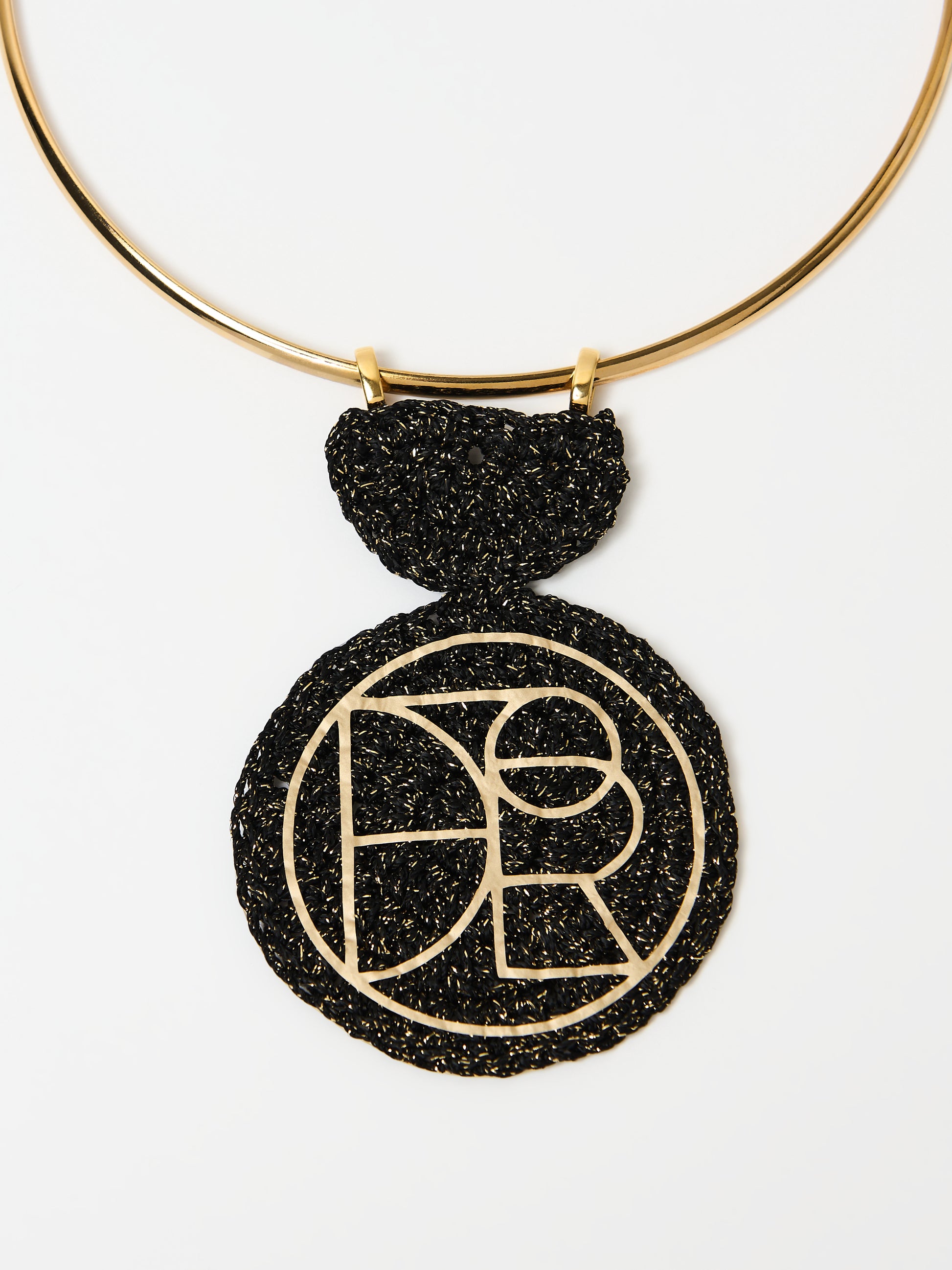 A close-up view of a gold choker necklace featuring a bold black textured pendant. The pendant is intricately crafted with a circular design, showcasing a gold geometric logo or pattern that contrasts elegantly against the black background. The detailed texture and gold accents highlight the luxurious and modern aesthetic of the piece.