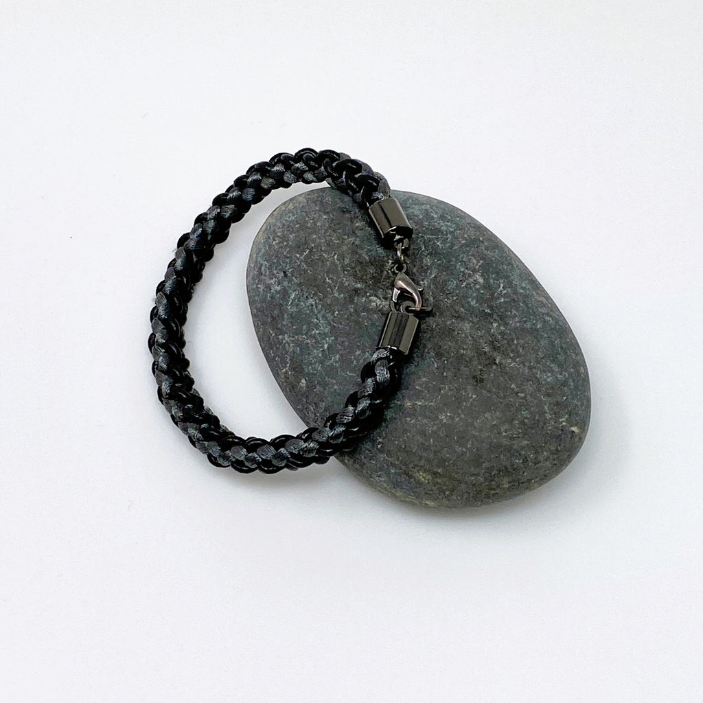 The Marlowe Midnight Black and Graphite Grey braided leather bracelet with a silver lobster clasp, laid flat, alternate view