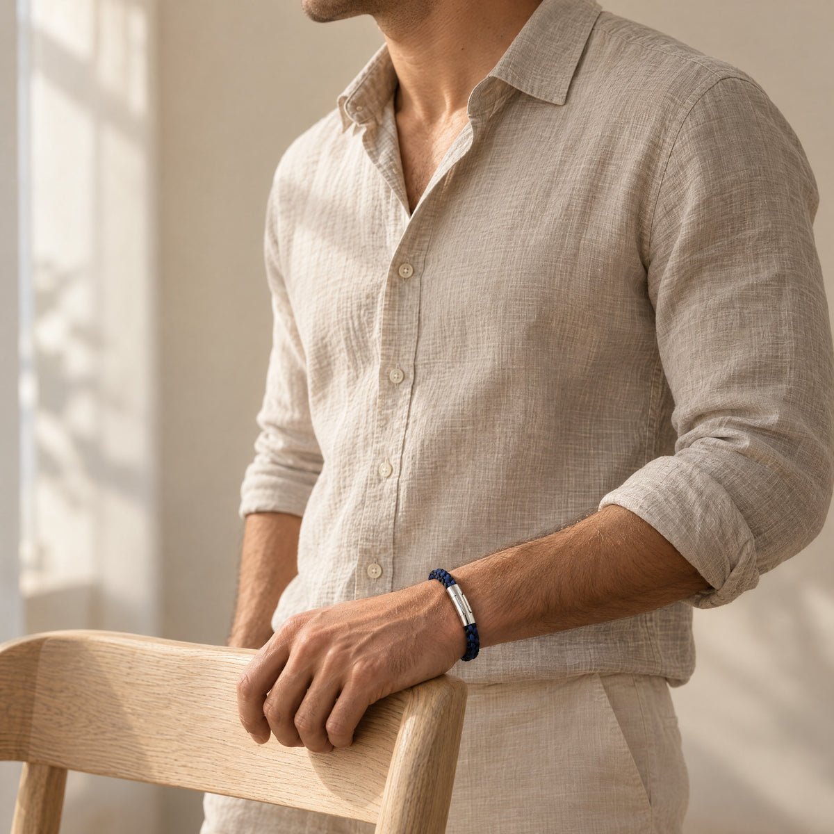 The Arden men's braided leather bracelet in Indigo Blue, worn on the wrist with one hand resting on a chair, polished silver magnetic clasp