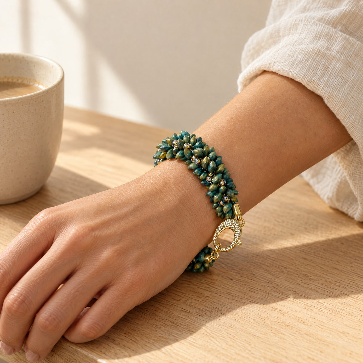 The Elio beaded bracelet for women in green and gold with a crystal-set gold lobster clasp, worn with the forearm resting on an oak table
