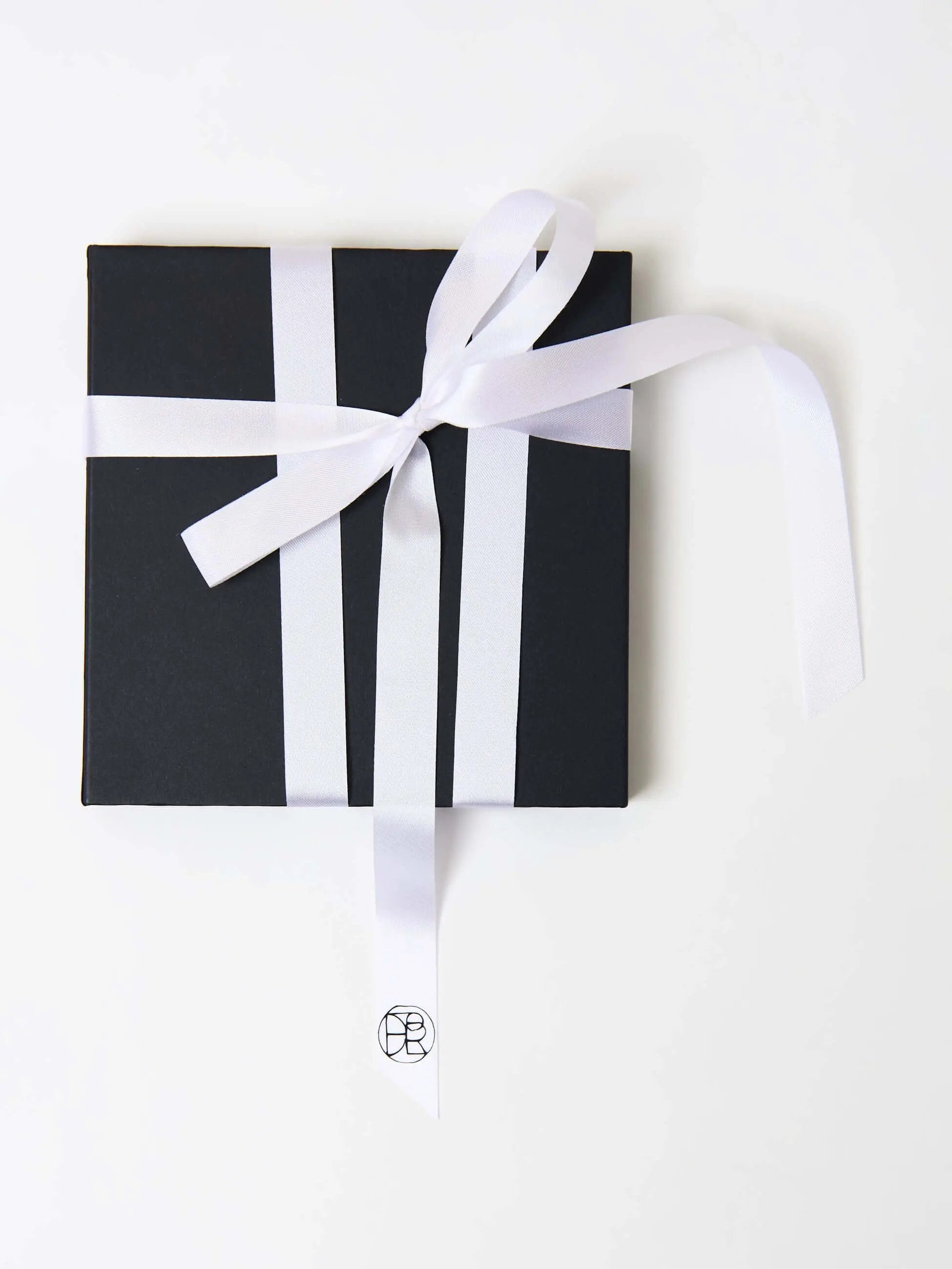 Black gift box with a neatly tied white ribbon in a cross pattern, featuring a minimalist logo on the ribbon tail, placed on a white background.