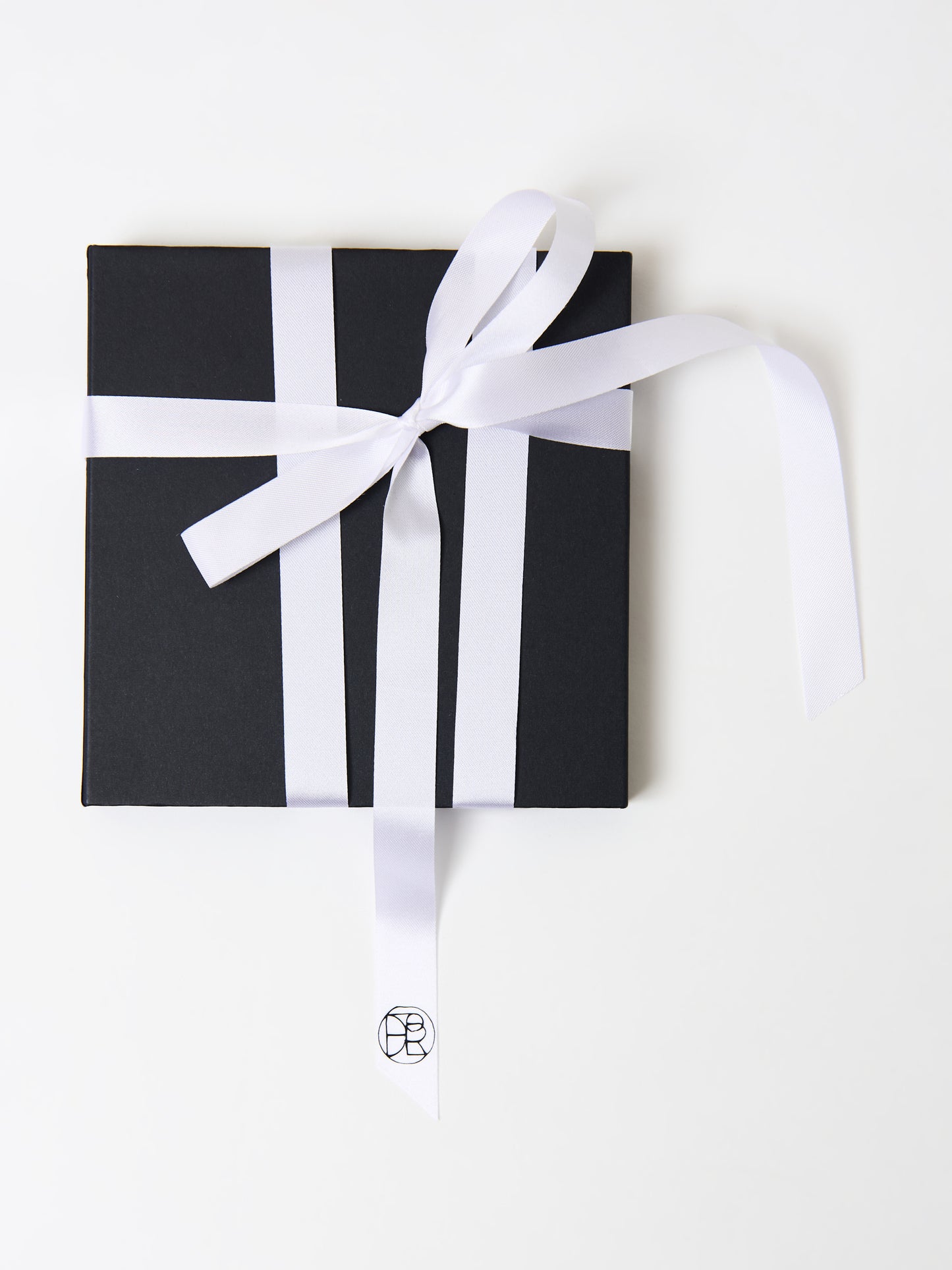 Black gift box with a neatly tied white ribbon in a cross pattern, featuring a minimalist logo on the ribbon tail, placed on a white background.