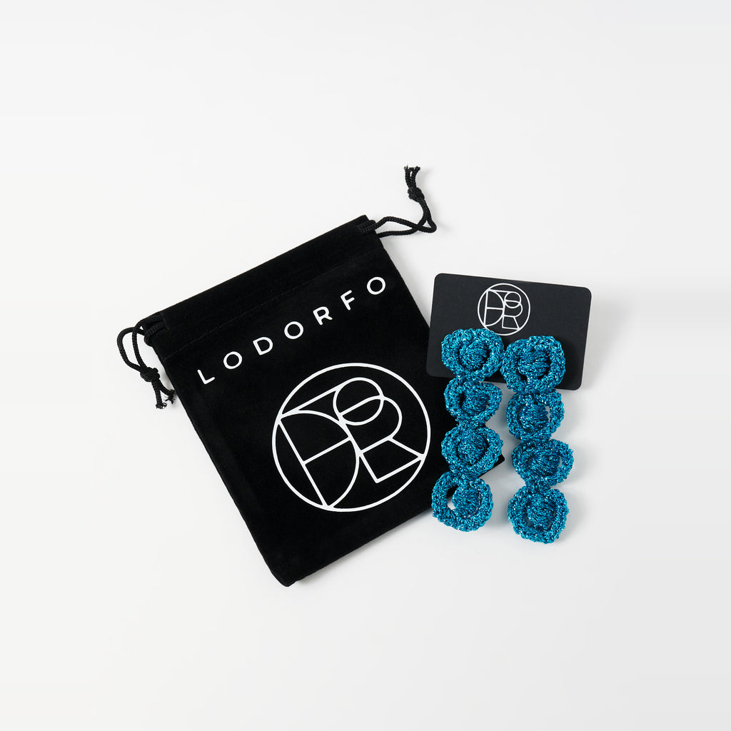 The Arlo long deep teal four-ring crochet loop earrings on a black earring card beside the black velvet gift pouch - Lodorfo