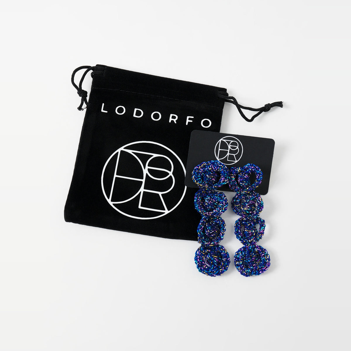 The Arlo long indigo blue four-ring crochet loop earrings on a black earring card beside the black velvet gift pouch - Lodorfo