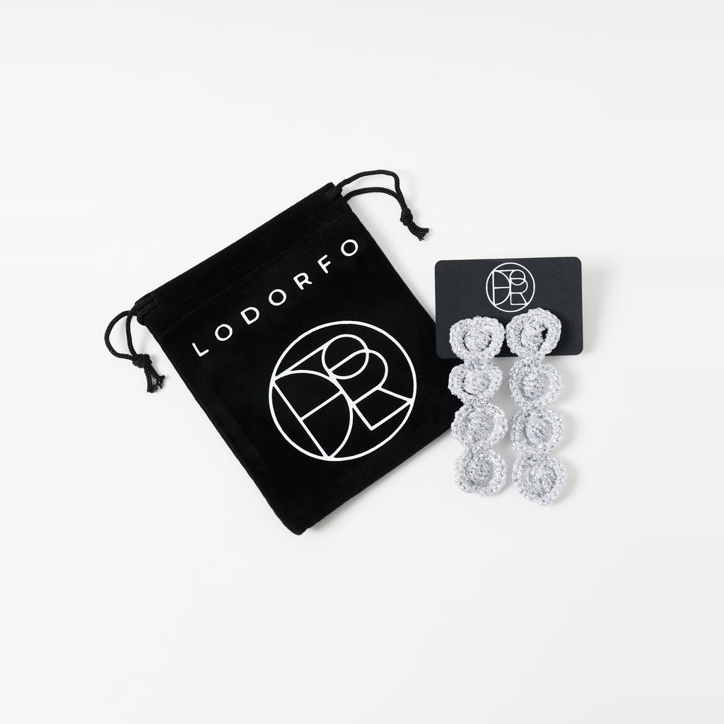 The Arlo long silver grey four-ring crochet loop earrings on a black earring card beside the black velvet gift pouch - Lodorfo