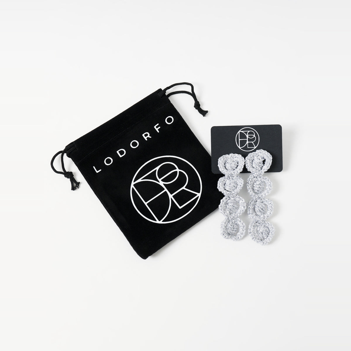 The Arlo long silver grey four-ring crochet loop earrings on a black earring card beside the black velvet gift pouch - Lodorfo