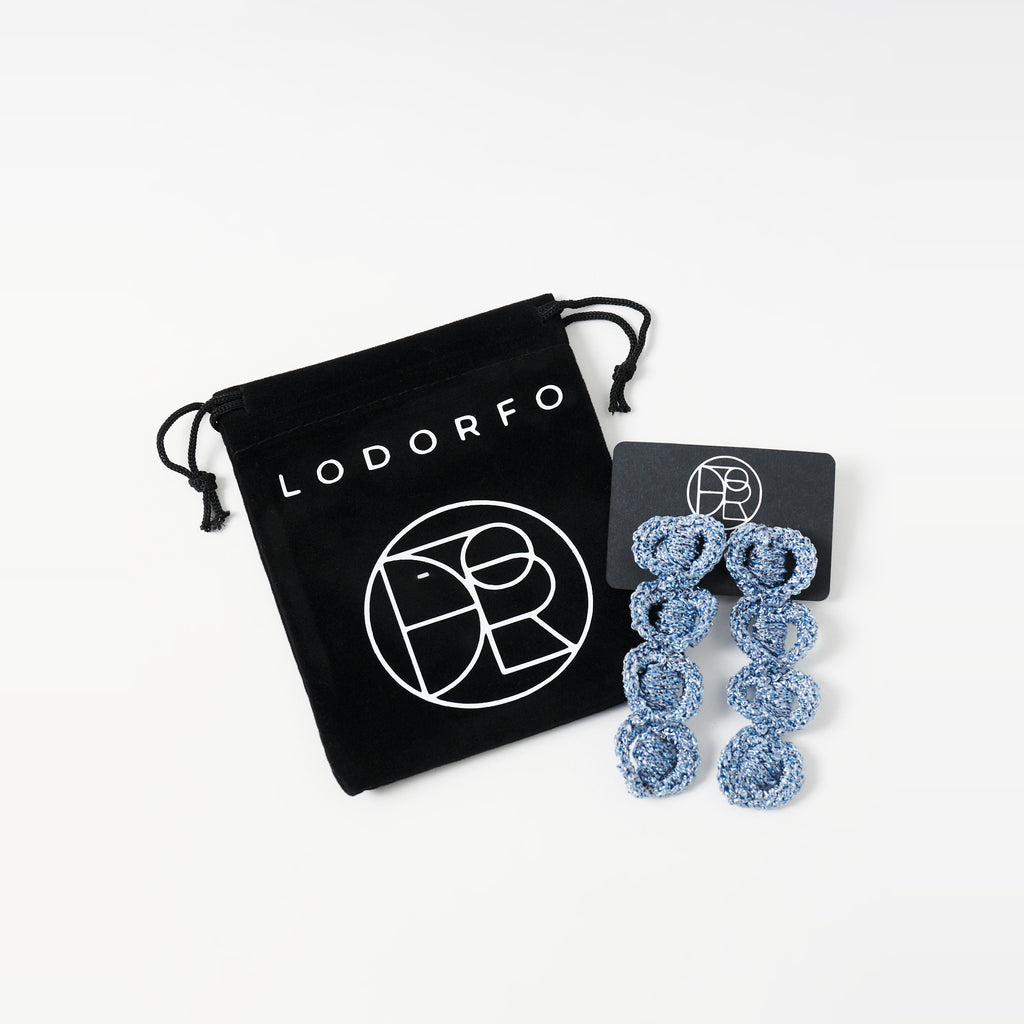 The Arlo long sky blue four-ring crochet loop earrings on a black earring card beside the black velvet gift pouch - Lodorfo