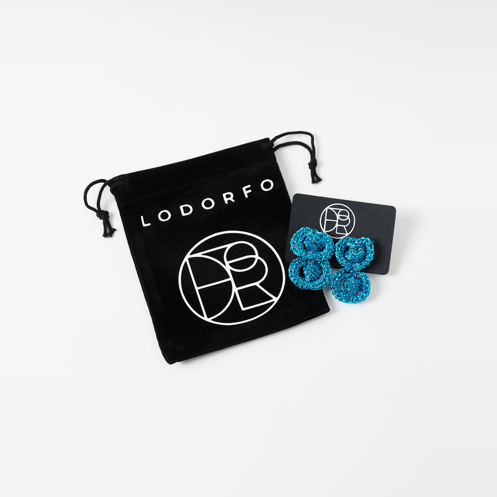 The Arlo short deep teal two-ring crochet loop earrings on a black earring card beside the black velvet gift pouch - Lodorfo