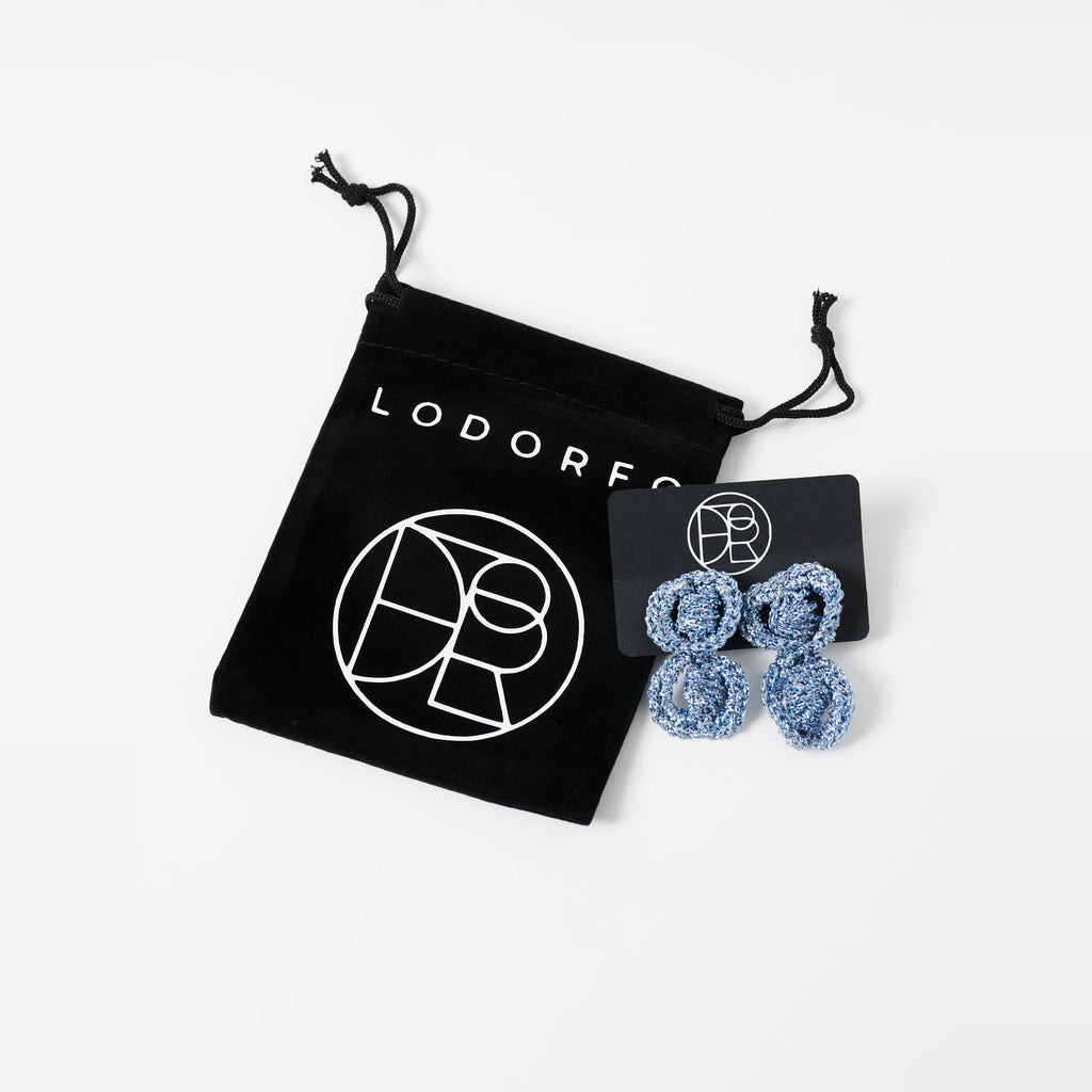 The Arlo short sky blue two-ring crochet loop earrings on a black earring card beside the black velvet gift pouch - Lodorfo