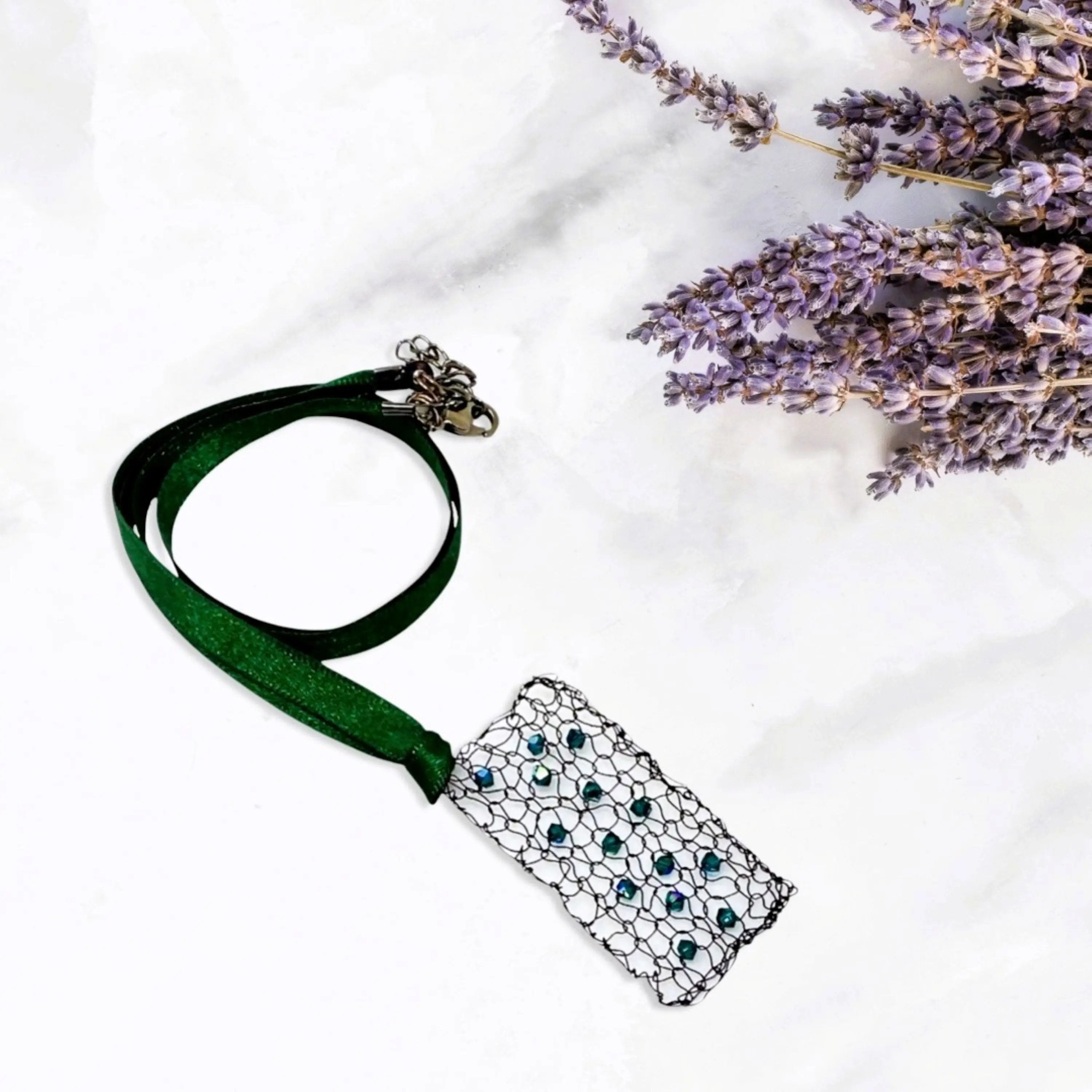 Flat lay of the necklace on a marble-like background with sprigs of dried lavender, featuring a green satin ribbon and a rectangular pendant woven with emerald crystal beads.