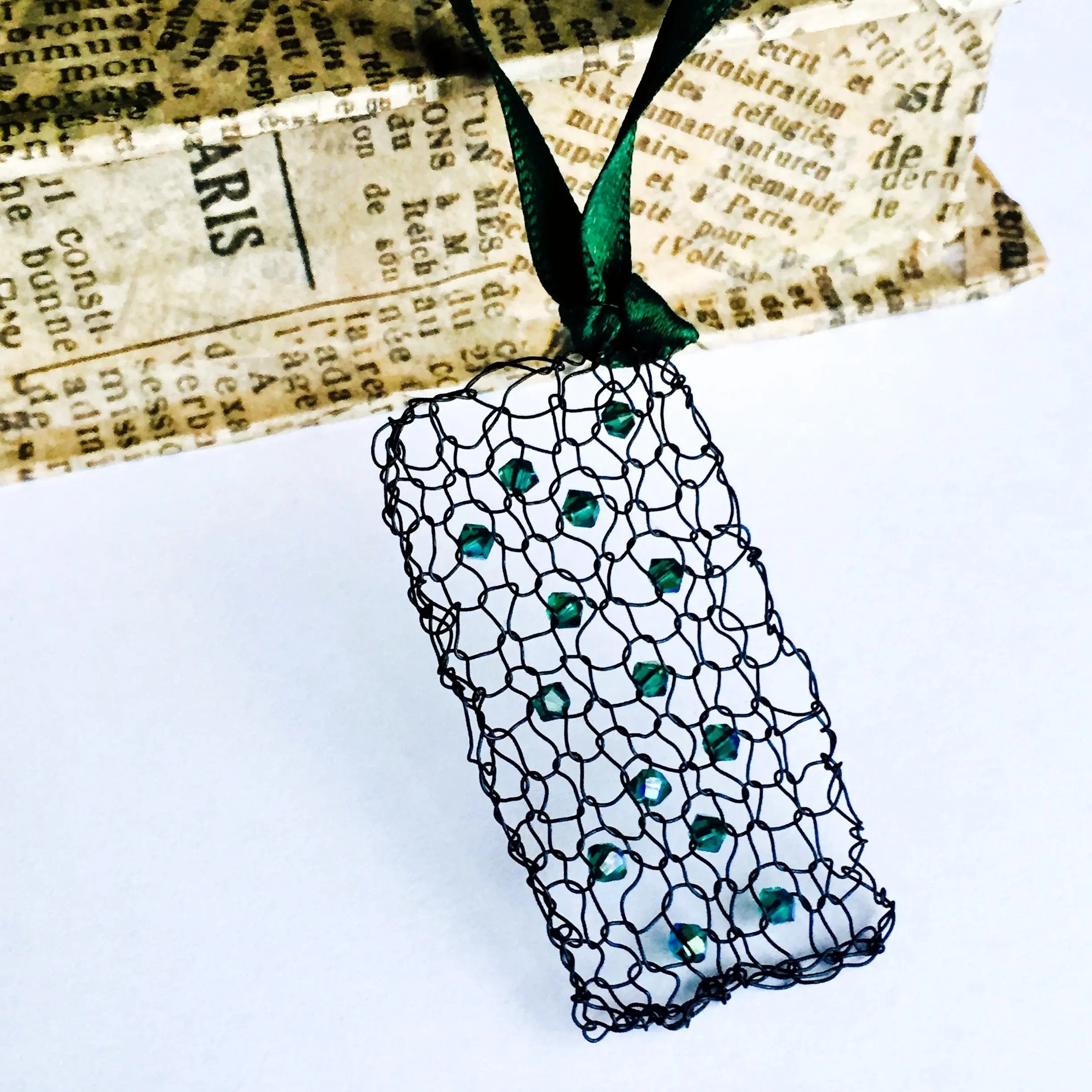 Handcrafted wire pendant necklace featuring emerald-green crystals woven into a rectangular black mesh, suspended from a green satin ribbon, styled against vintage newspaper packaging.