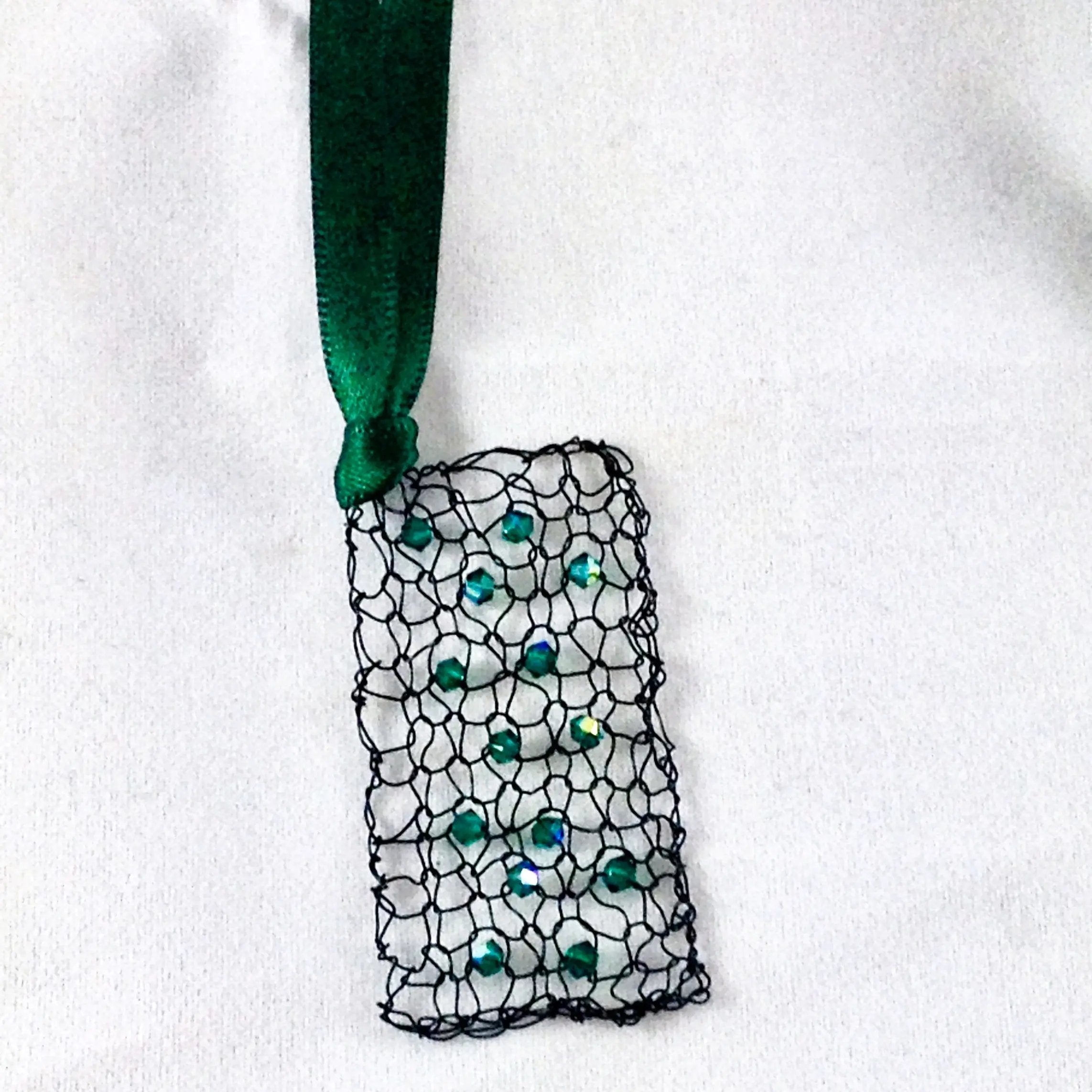 Emerald crystal wire mesh pendant hanging vertically against a white fabric background, showcasing its delicate lace-like structure and green ribbon attachment.