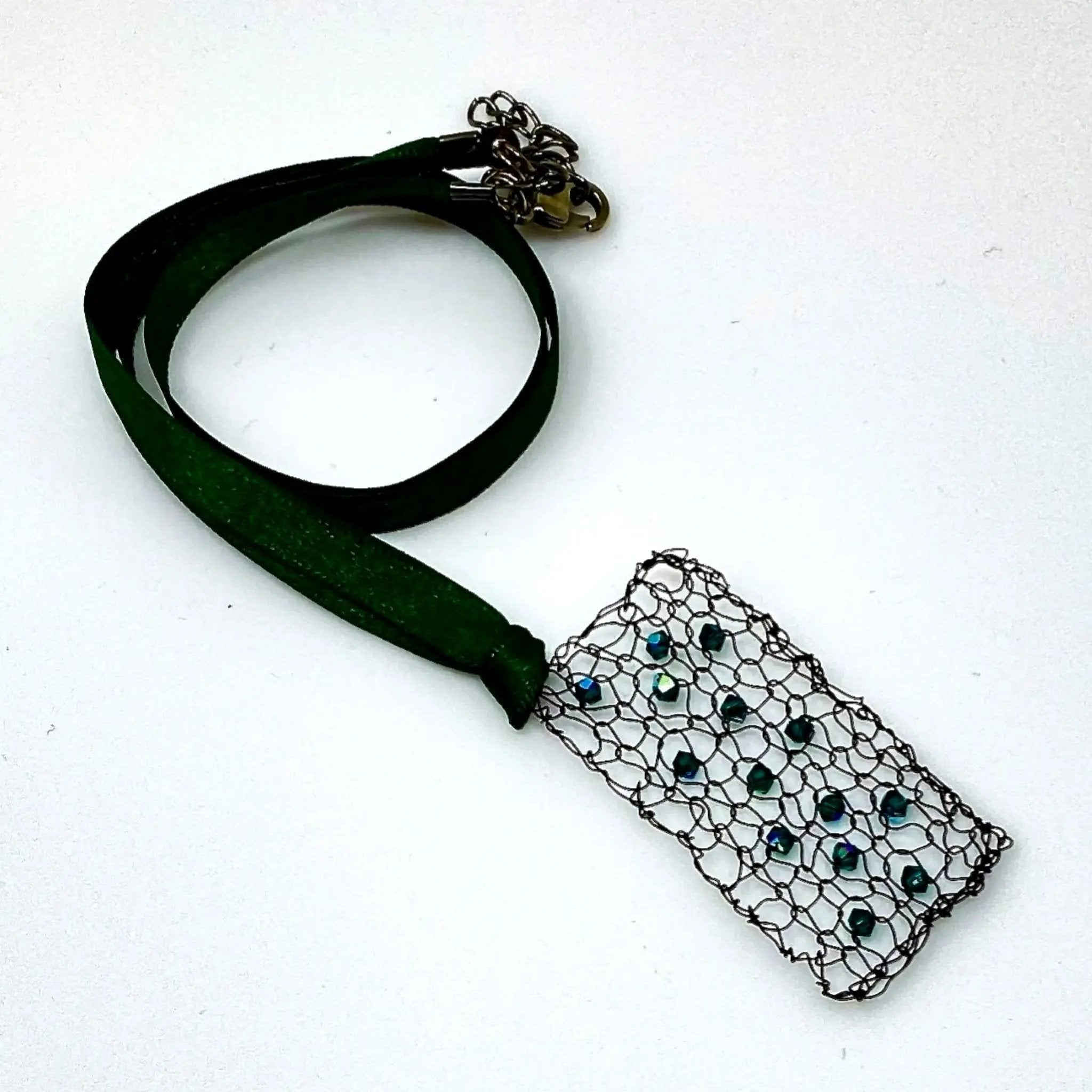 Full necklace view showing the green satin ribbon and clasp paired with a rectangular pendant made of black wire mesh and emerald-coloured crystals.