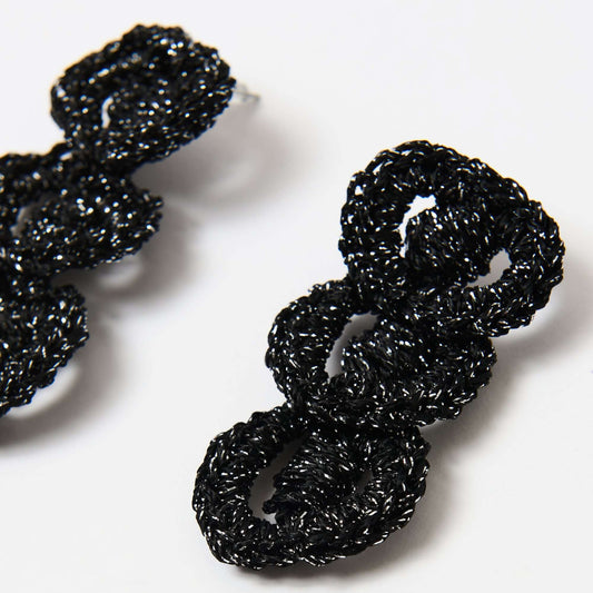 Close-up of handcrafted black crochet loop earrings with a glossy, metallic sheen, featuring interlocking circular shapes on a white background.