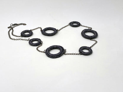 A statement necklace featuring textured dark metallic circular links of varying sizes, connected by a delicate chain. The unique design showcases an industrial-chic aesthetic, perfect for bold and contemporary fashion styles.