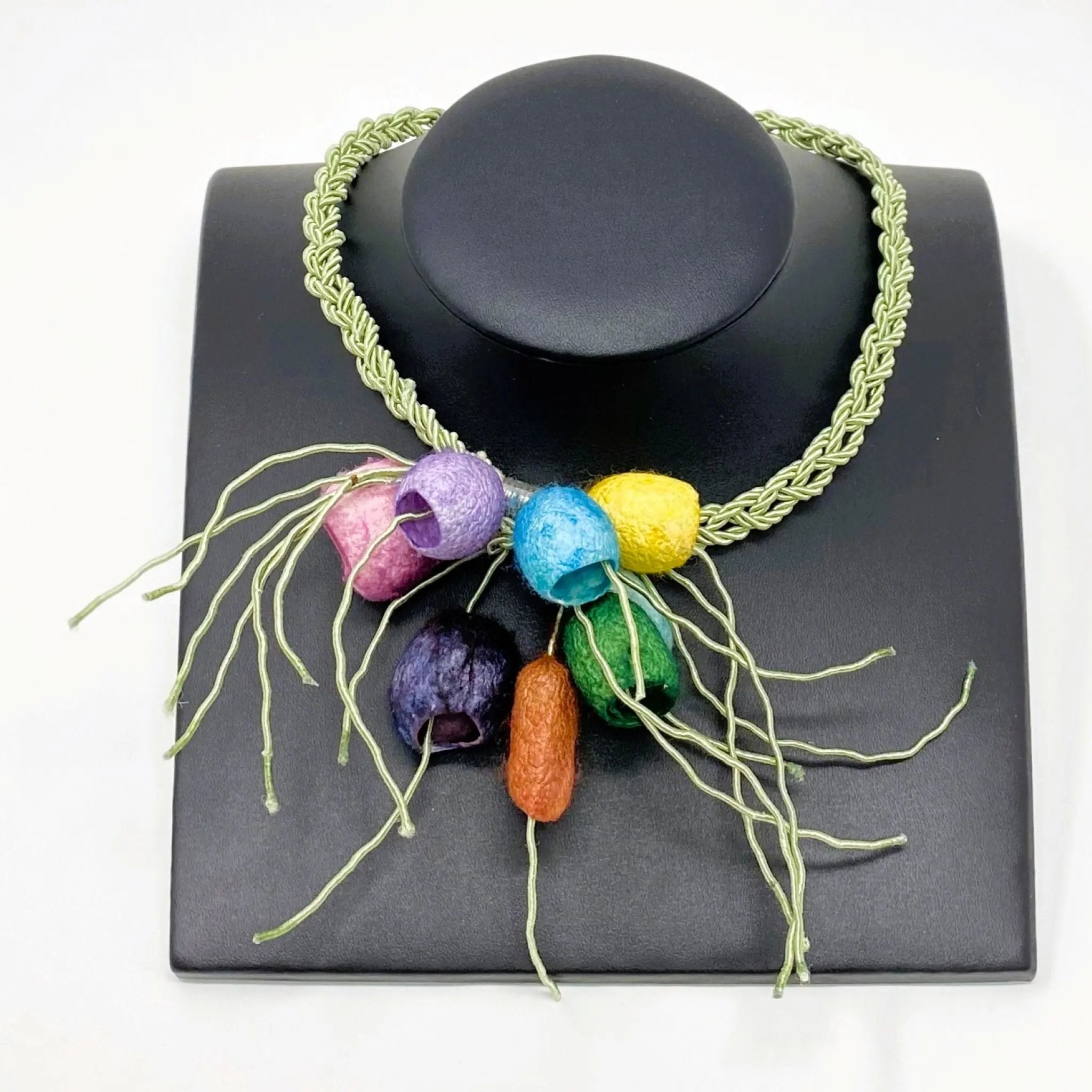 Handcrafted statement necklace featuring a braided green cord with a cluster of silk cocoons displayed on a leather bust..