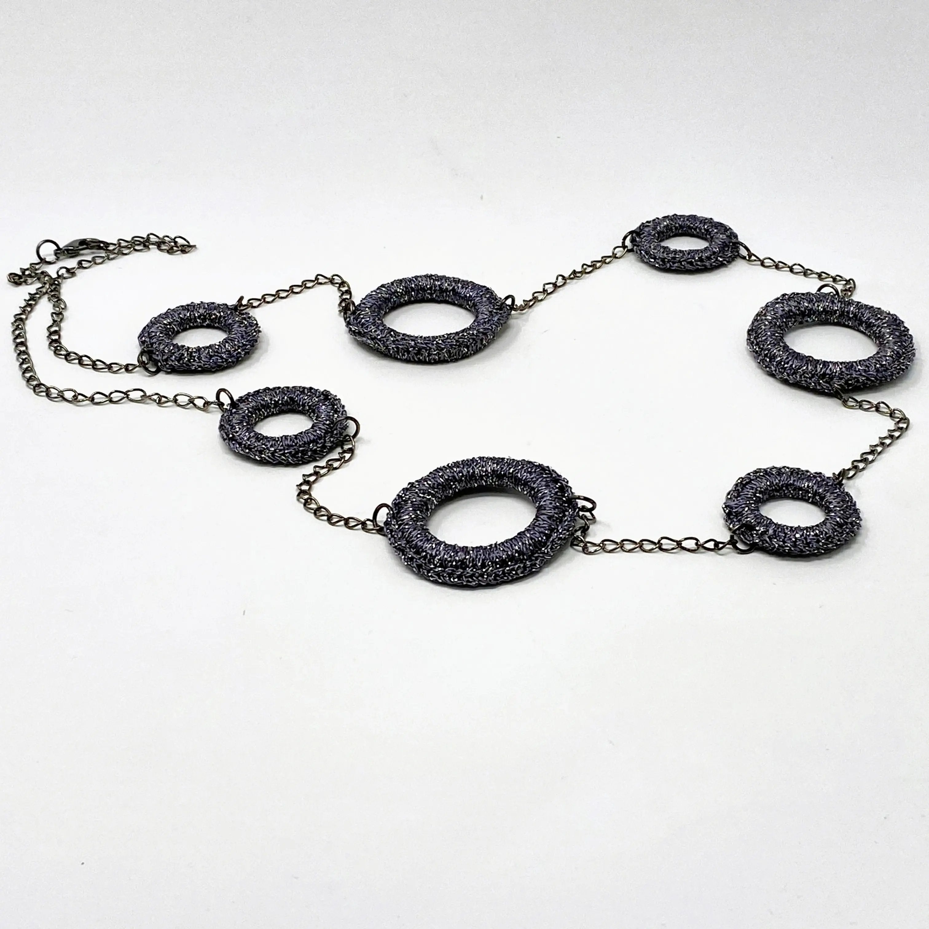 Long necklace composed of shimmering charcoal-coloured ring elements connected by dark chain links, laid flat against a white background.