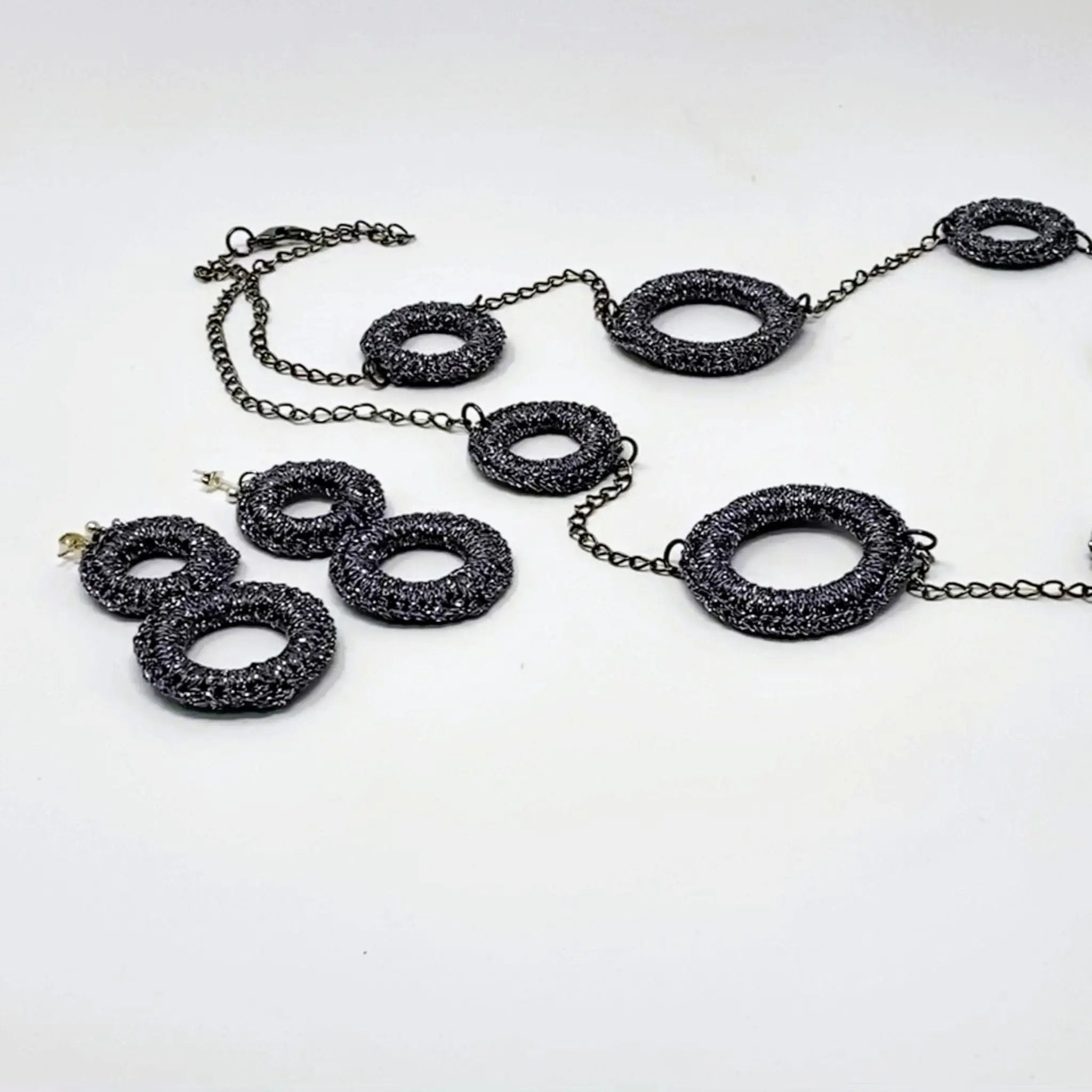 Handcrafted jewellery set featuring a long necklace and matching earrings, each adorned with textured charcoal-toned circular rings linked by a dark metal chain.