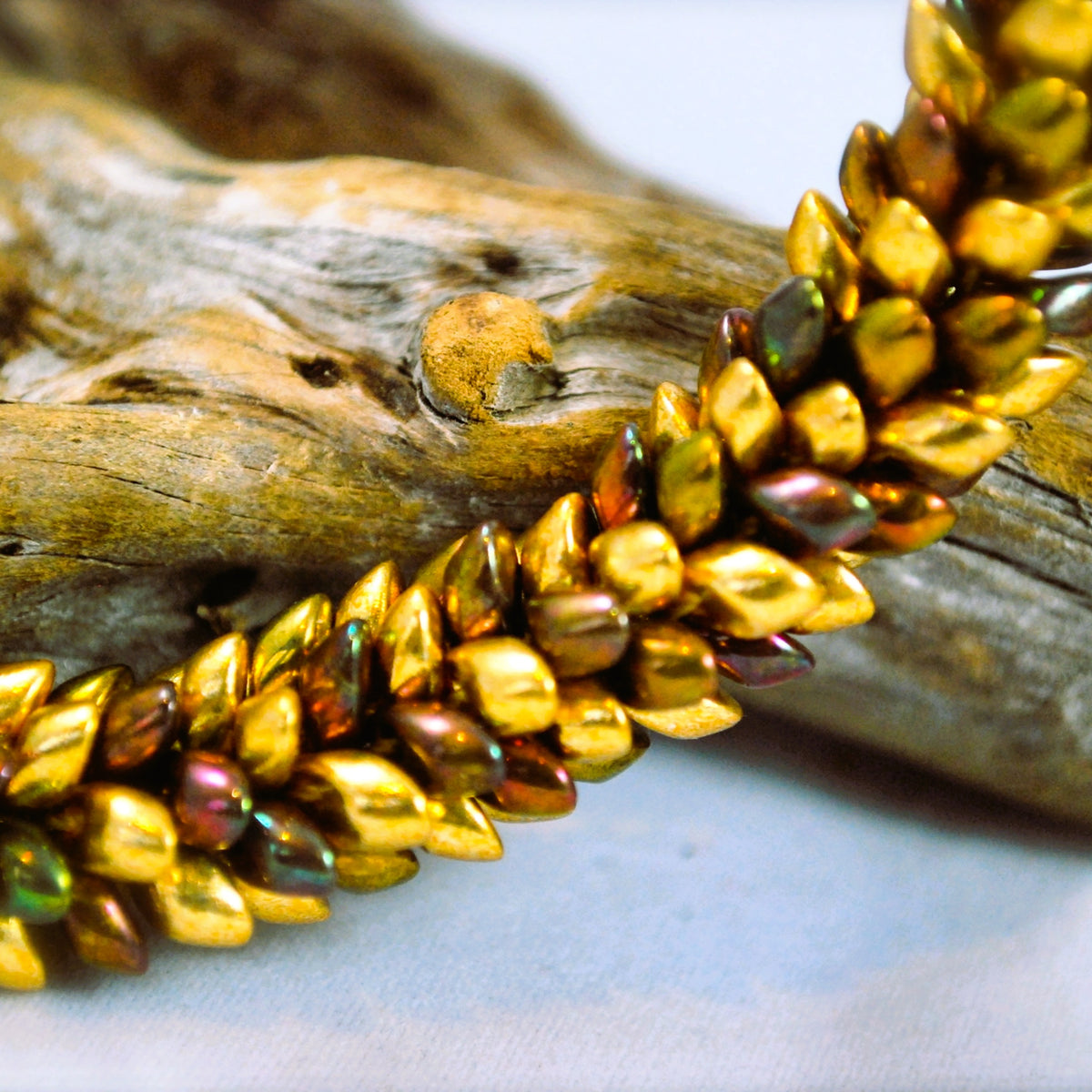 Close-up of the Maris braided magatama beadwork in gold, olive and bronze tones resting on driftwood - Lodorfo