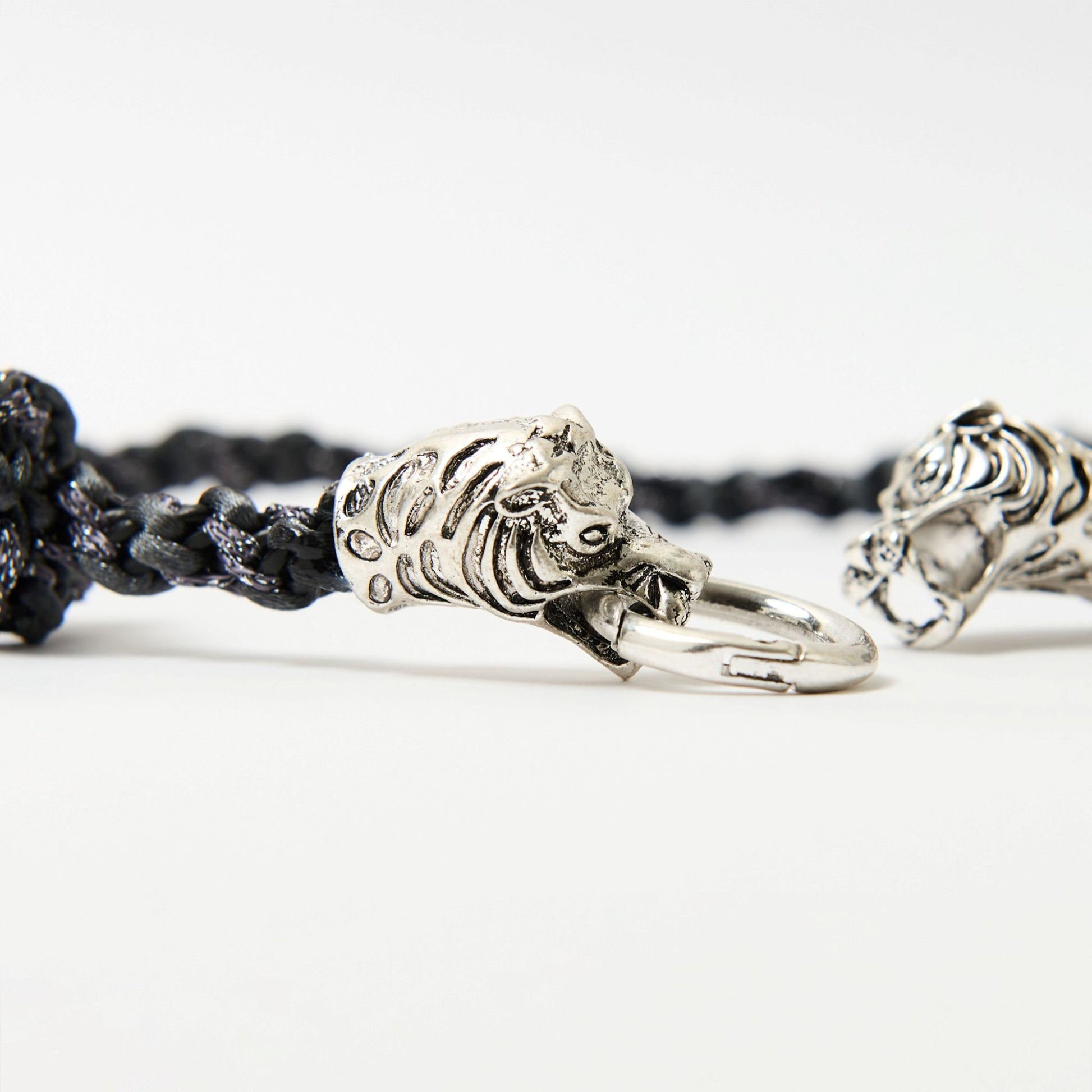 Close-up of a black braided cord necklace featuring a silver tiger head clasp with intricate detailing, each head gripping a central metal ring.