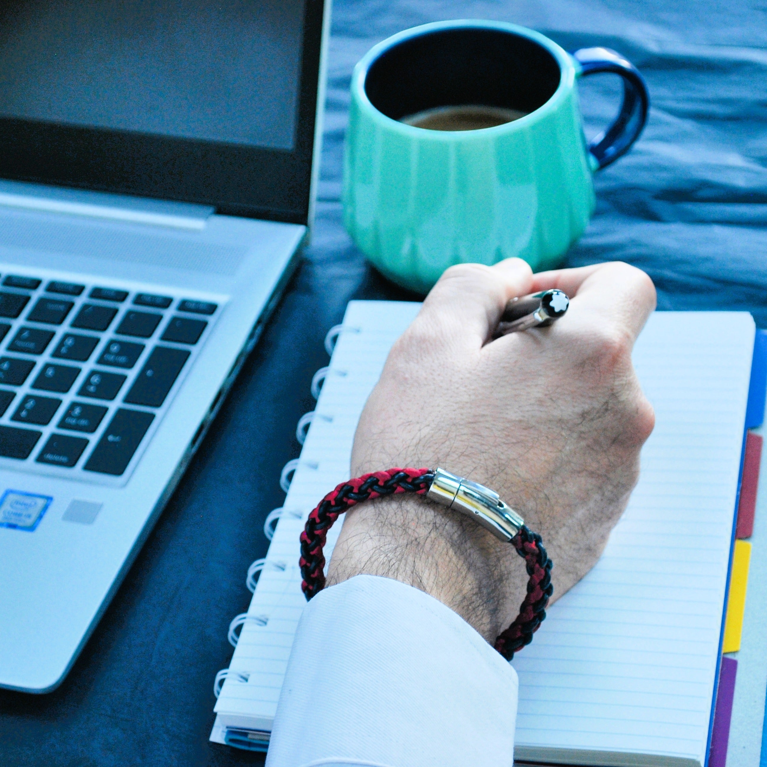 Person writing in a notebook with a teal mug and laptop on a table