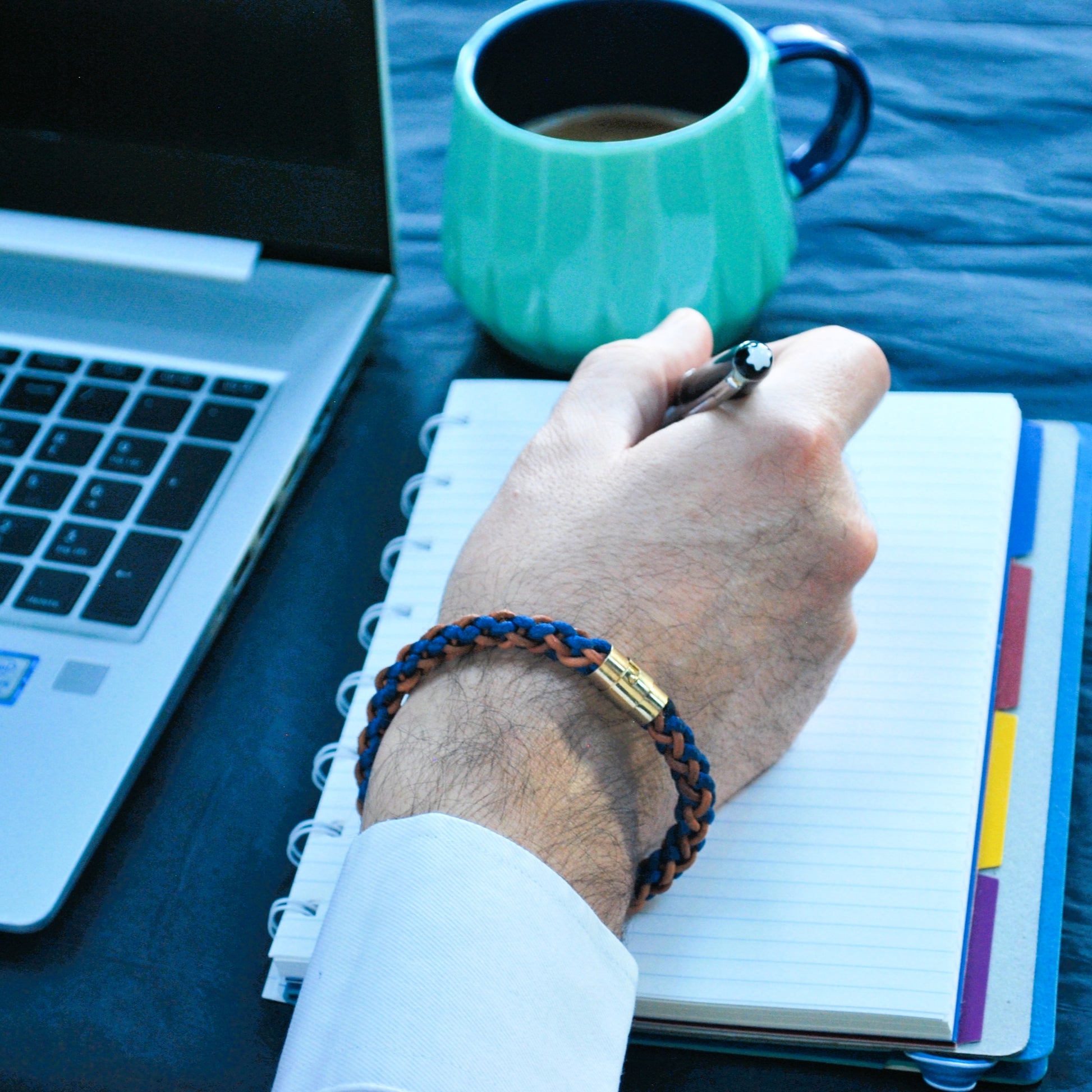 Lifestyle image of a men's blue and brown braided leather bracelet with gold magnetic clasp, shown on wrist while writing in notebook — artisan accessory for modern professionals.