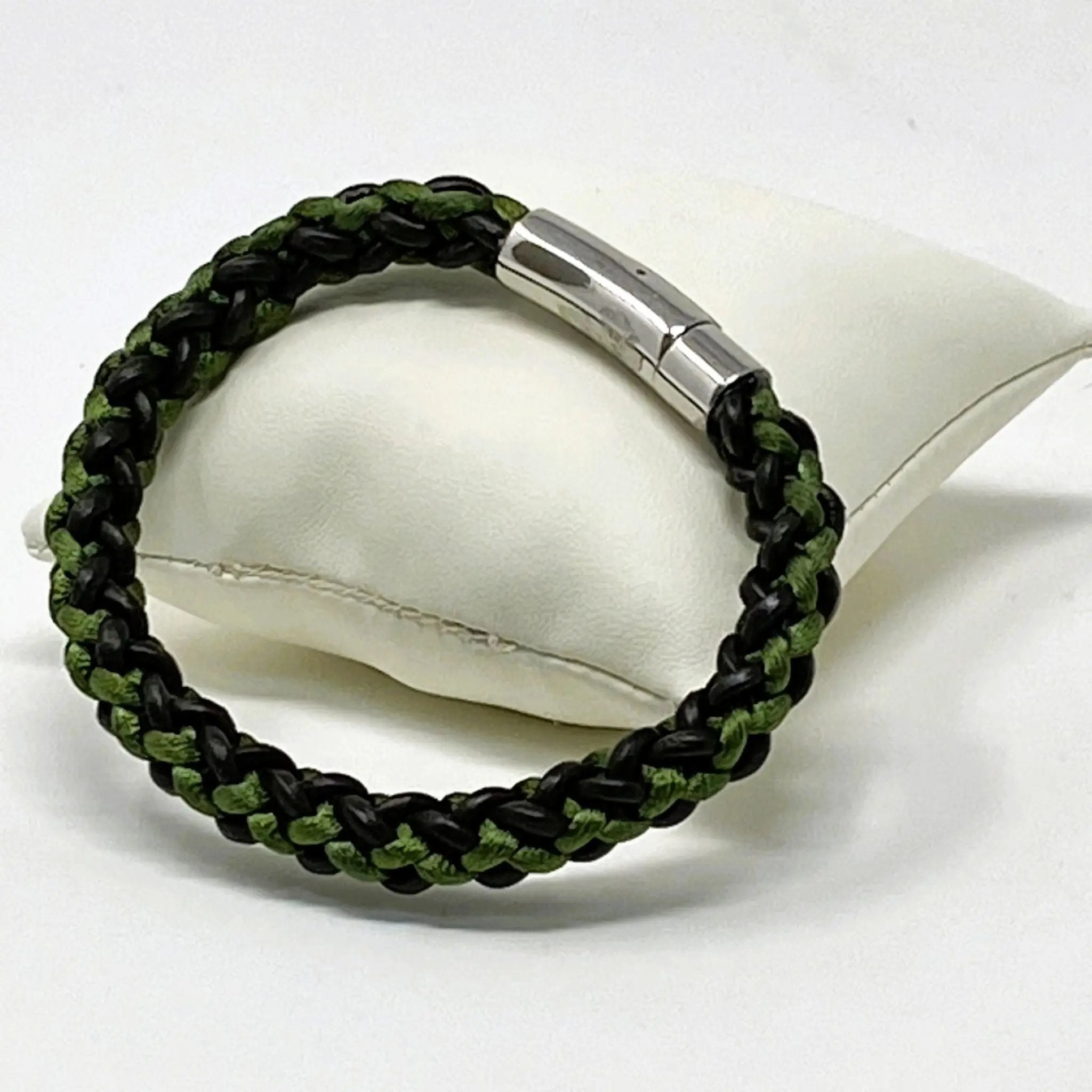 Green and brown braided leather bracelet for men, shown on a white display cushion with polished stainless steel clasp — rugged yet refined accessory for casual wear.