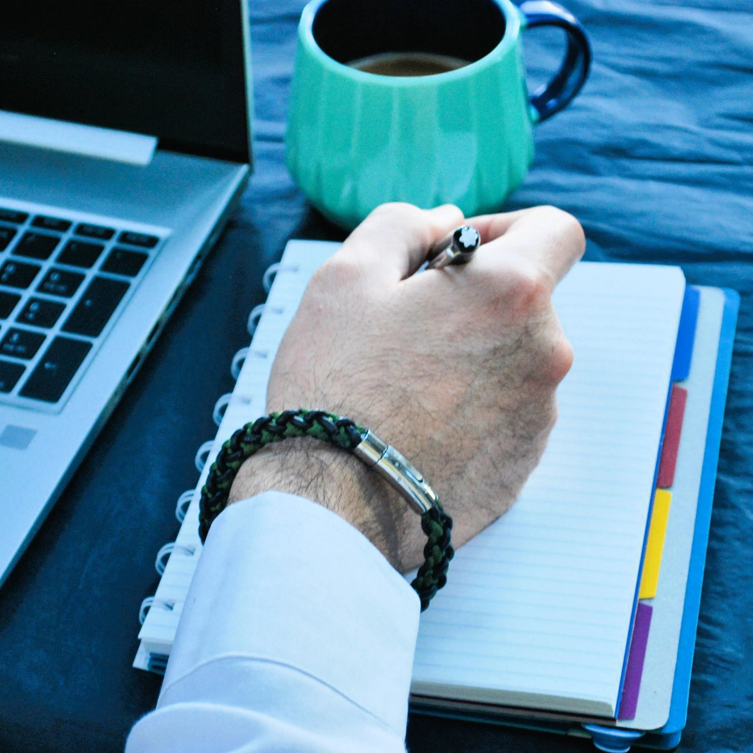 Lifestyle photo of men’s green and brown braided leather bracelet with polished silver clasp, shown on wrist while journaling at a desk — sleek, professional accessory for everyday use.