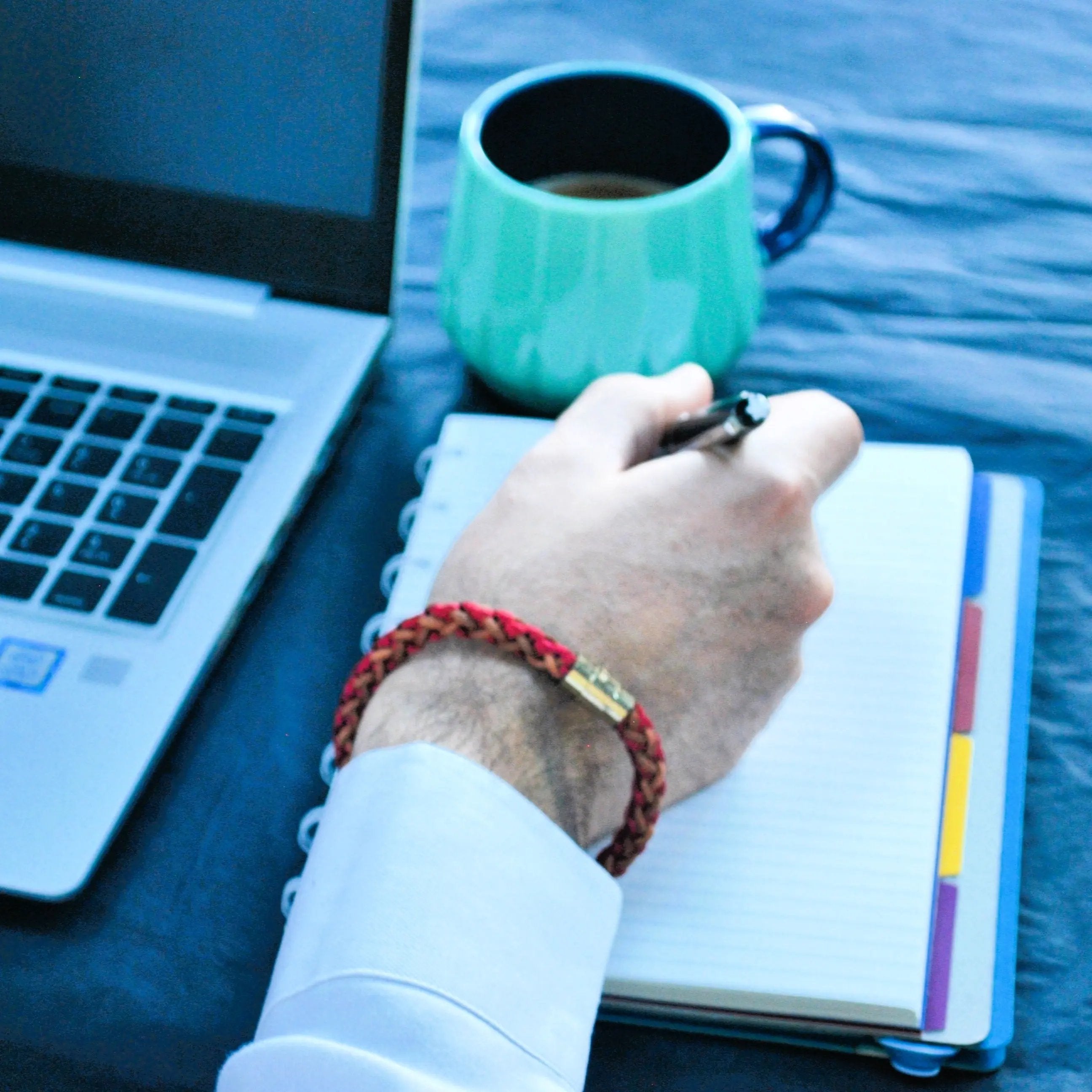 Lifestyle image of a men's red and brown braided leather bracelet with gold magnetic clasp, shown on wrist while writing in notebook — artisan accessory for modern professionals.