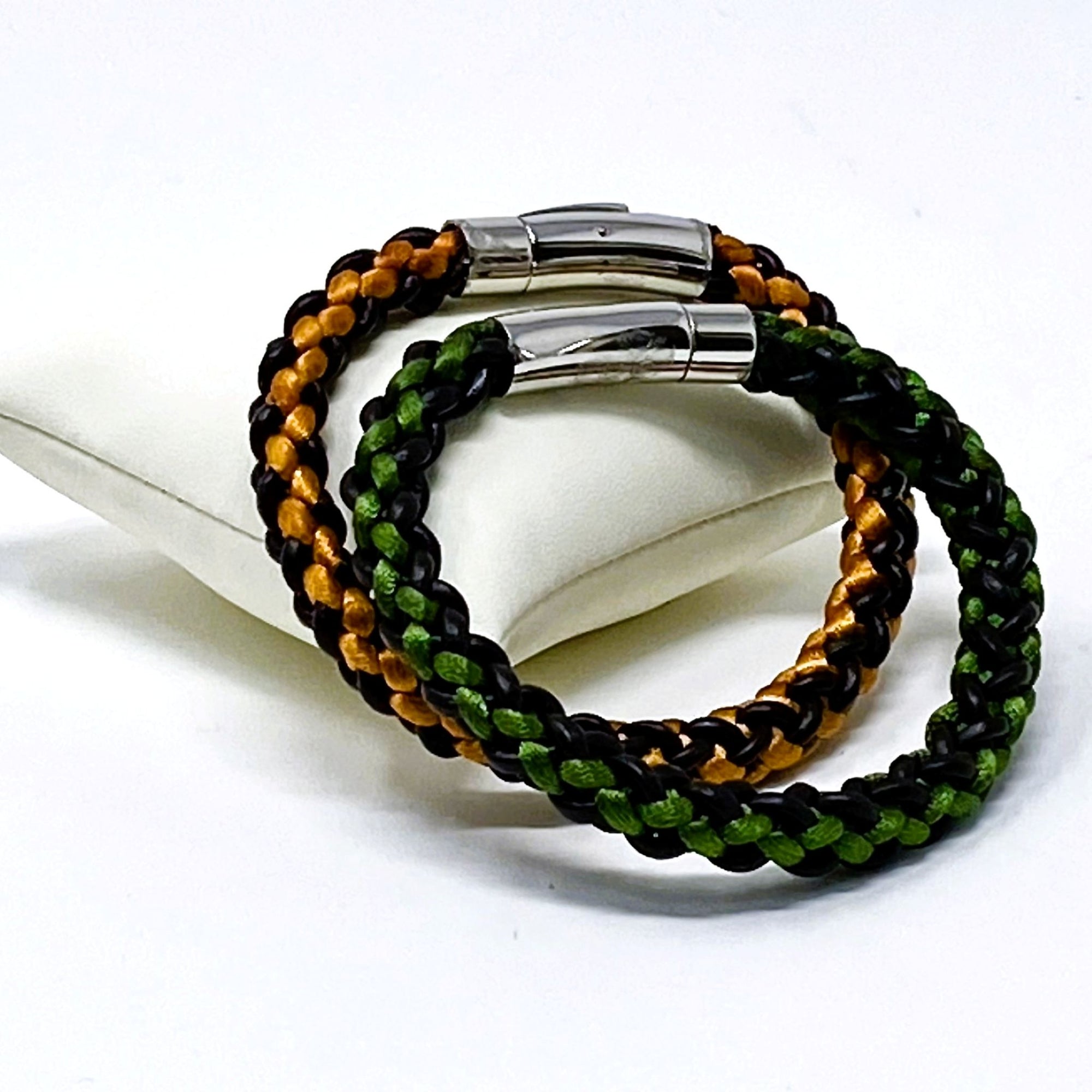 Green and orange braided leather bracelets for men, styled on a white display cushion with polished stainless steel clasps — artisan-crafted accessories perfect for gifting or matching.