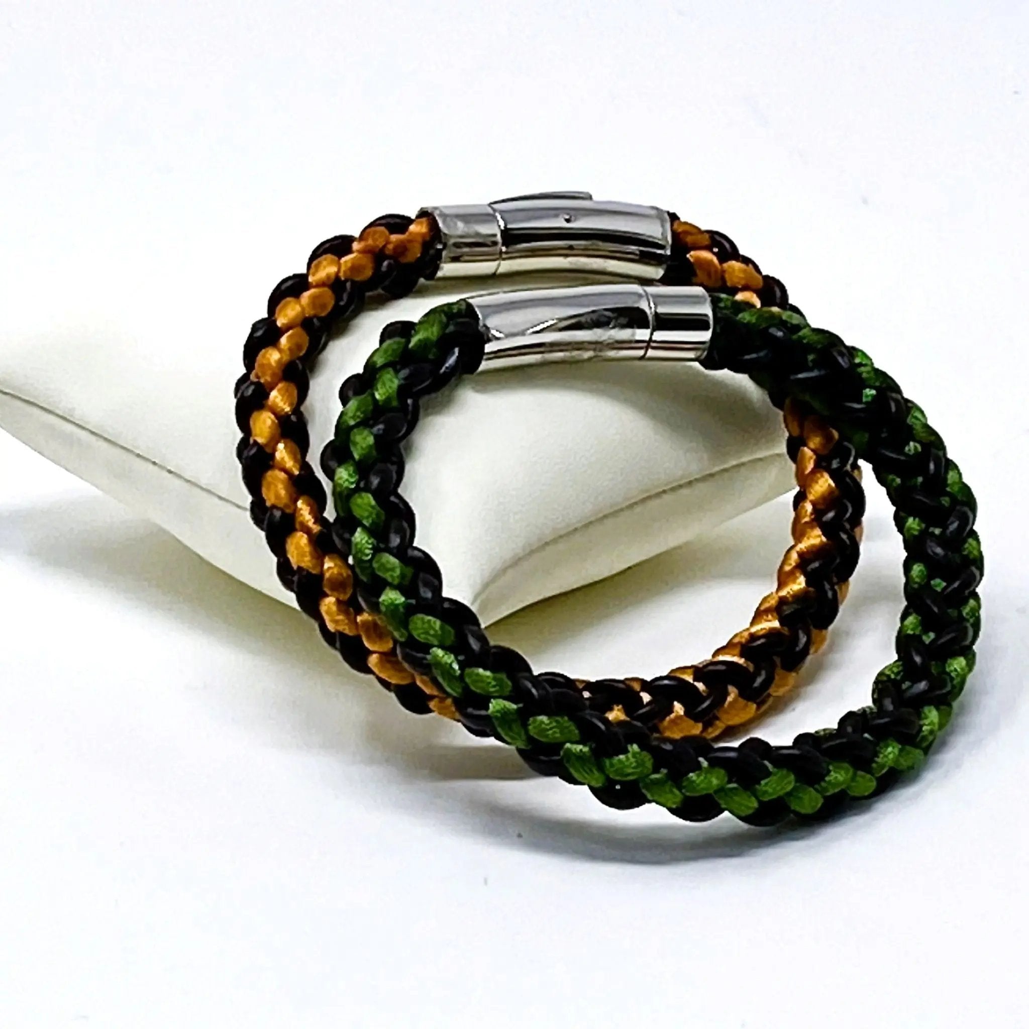 Green and orange braided leather bracelets for men, styled on a white display cushion with polished stainless steel clasps — artisan-crafted accessories perfect for gifting or matching.