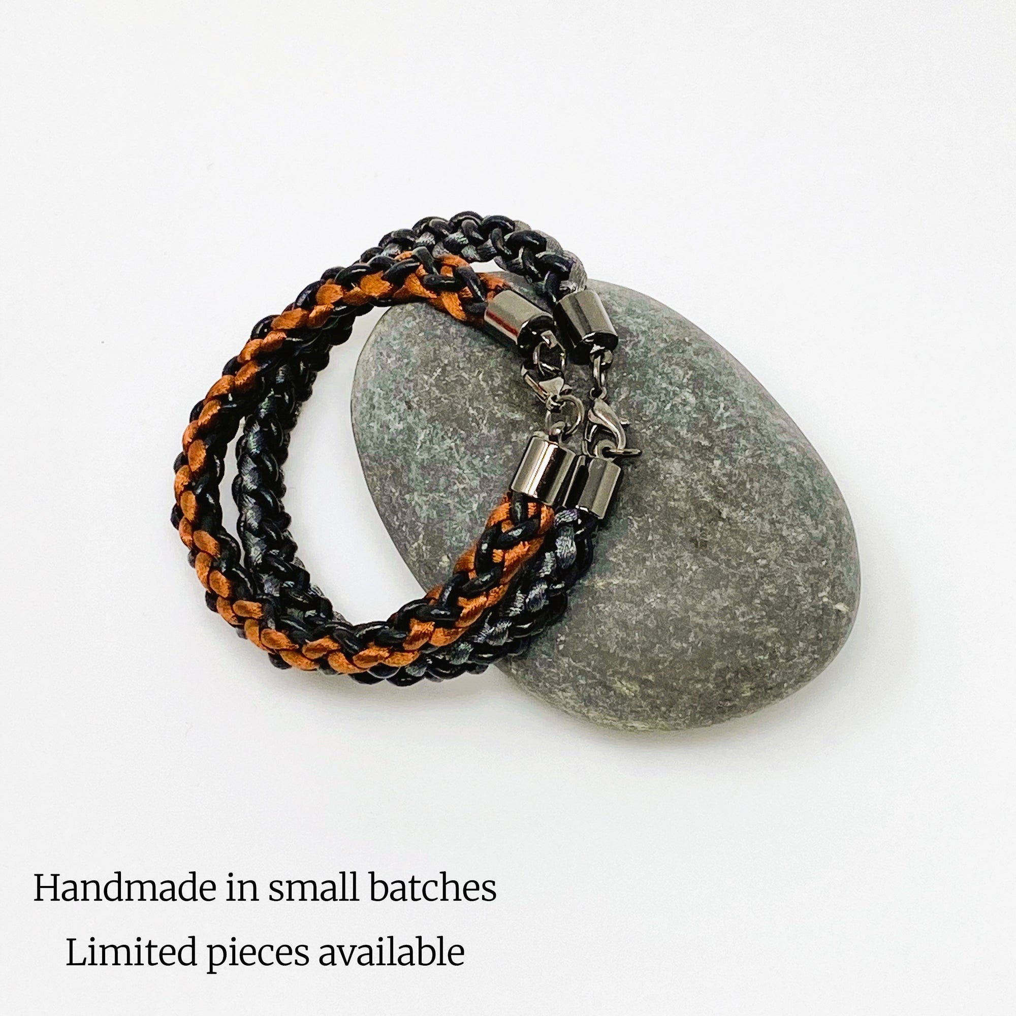 Artisan-crafted braided bracelets in black with orange and grey accents, styled on a natural stone — rugged men’s accessory duo with sleek gunmetal clasps.