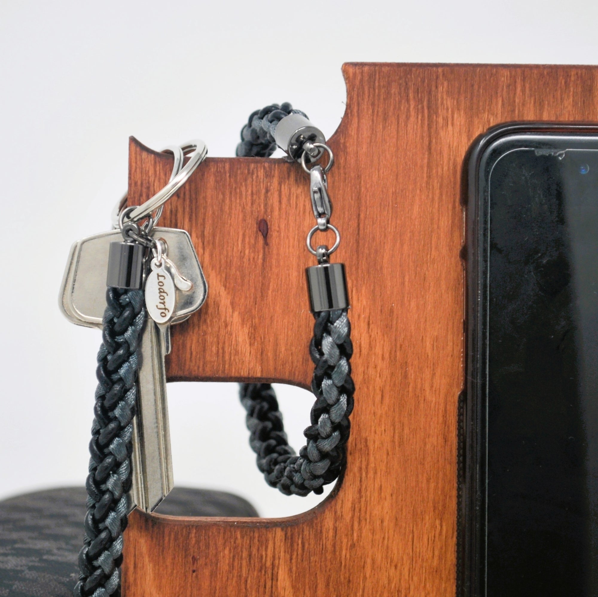 Lifestyle shot of braided grey and black leather bracelet with sleek gunmetal clasp, hanging on a wood docking station with keys and smartphone — stylish men’s accessory meets daily function.