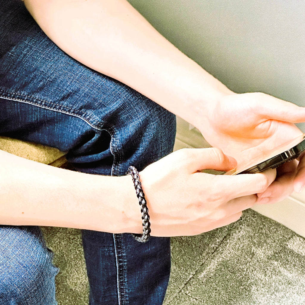 Lifestyle image of a man in jeans wearing a grey and black braided leather bracelet, shown while seated and holding a smartphone — casual yet refined wristwear for daily style.