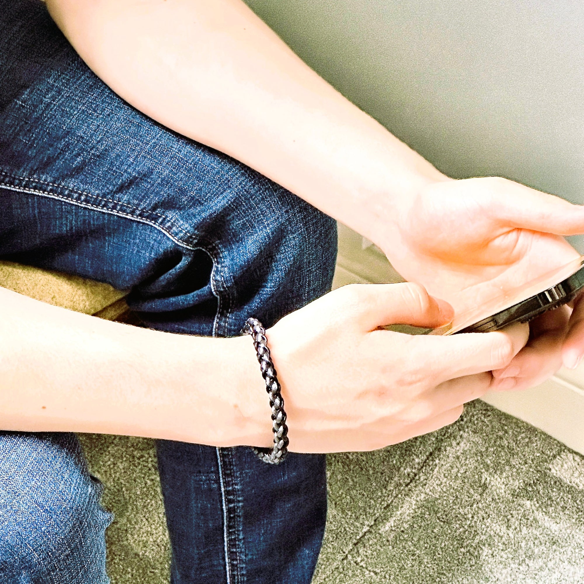 Lifestyle image of a man in jeans wearing a grey and black braided leather bracelet, shown while seated and holding a smartphone — casual yet refined wristwear for daily style.