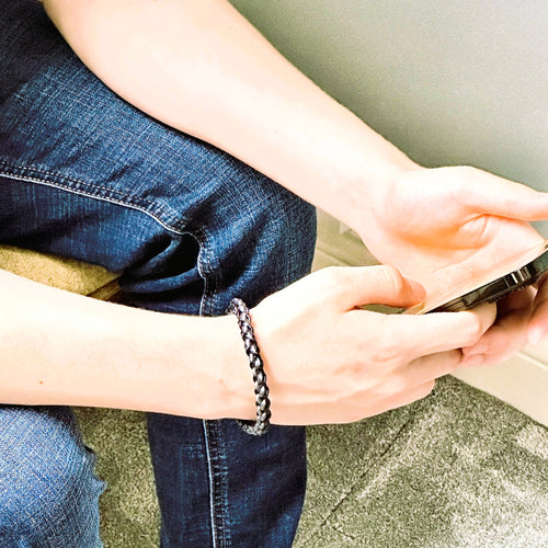 Lifestyle image of a man in jeans wearing a grey and black braided leather bracelet, shown while seated and holding a smartphone — casual yet refined wristwear for daily style.