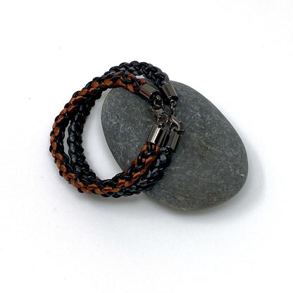 Two braided bracelets, one featuring brown and black tones and the other grey and black, both with stainless steel lobster clasps, elegantly arranged on a grey stone against a white background.