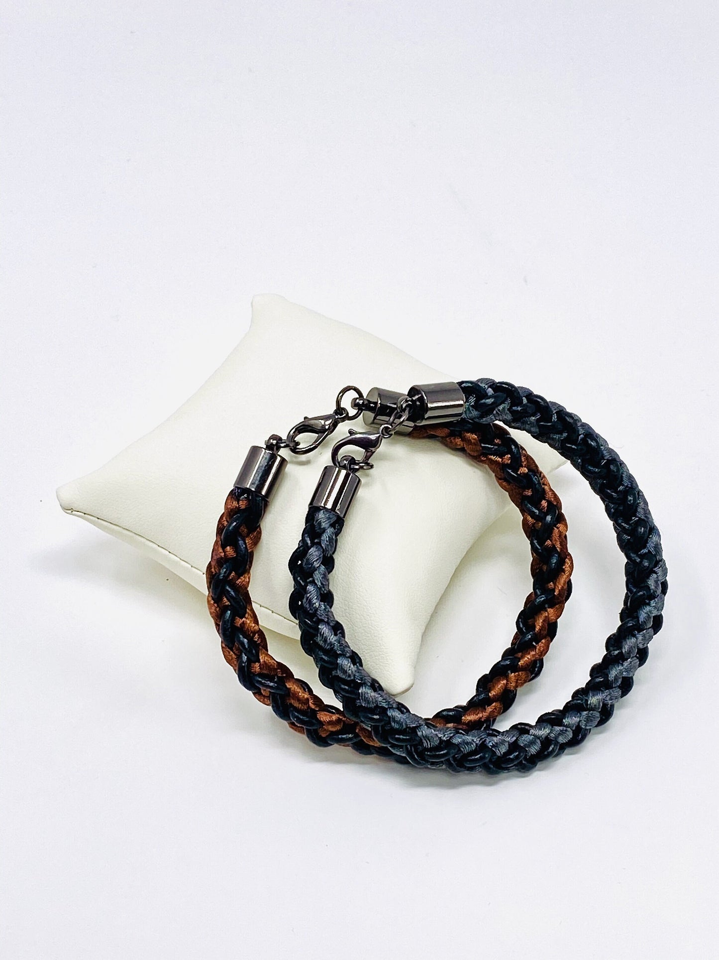 Two braided bracelets, one featuring brown and black tones and the other grey and black, both with stainless steel lobster clasps, elegantly arranged on an ivory leather cushion against a white background.