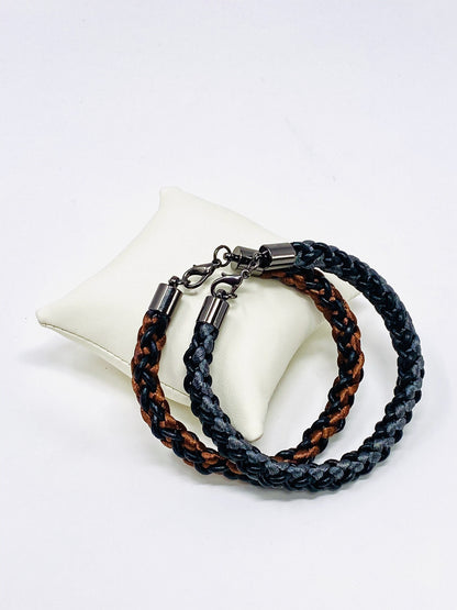 Two braided bracelets, one featuring brown and black tones and the other grey and black, both with stainless steel lobster clasps, elegantly arranged on an ivory leather cushion against a white background.