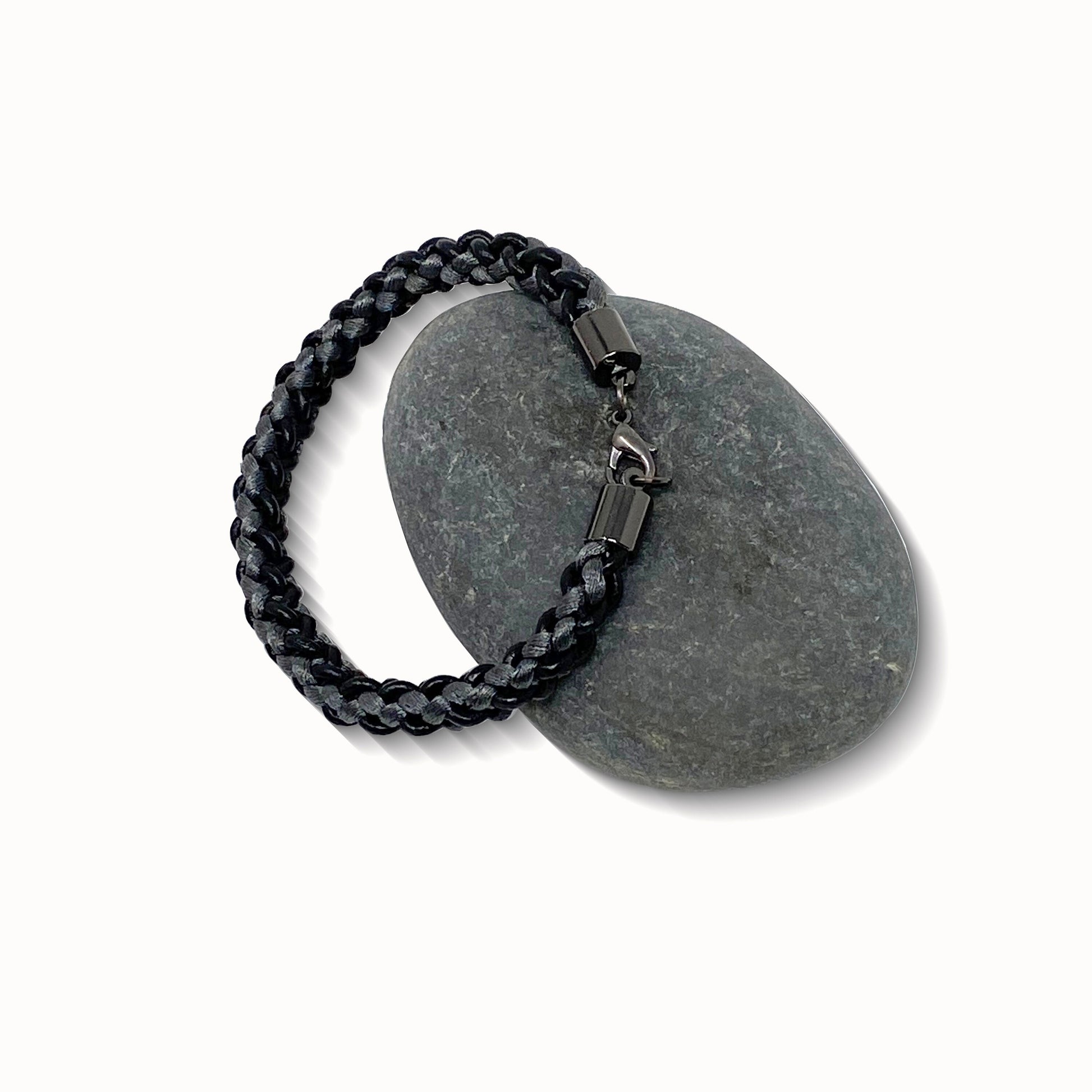 A braided bracelet with grey and black tones, featuring a stainless steel lobster clasp, elegantly displayed on a smooth stone against a white background.