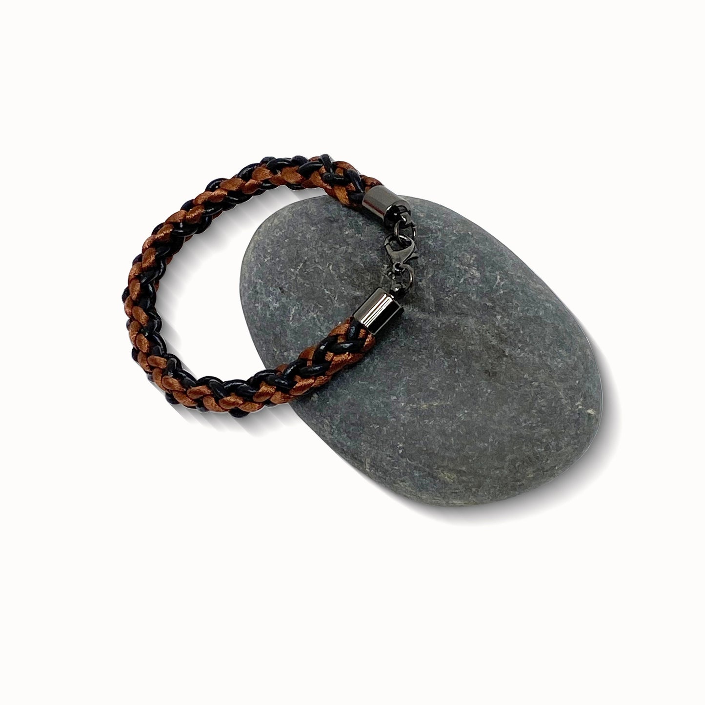 A braided bracelet with brown and black tones, featuring a stainless steel lobster clasp, elegantly displayed on a smooth stone against a white background.