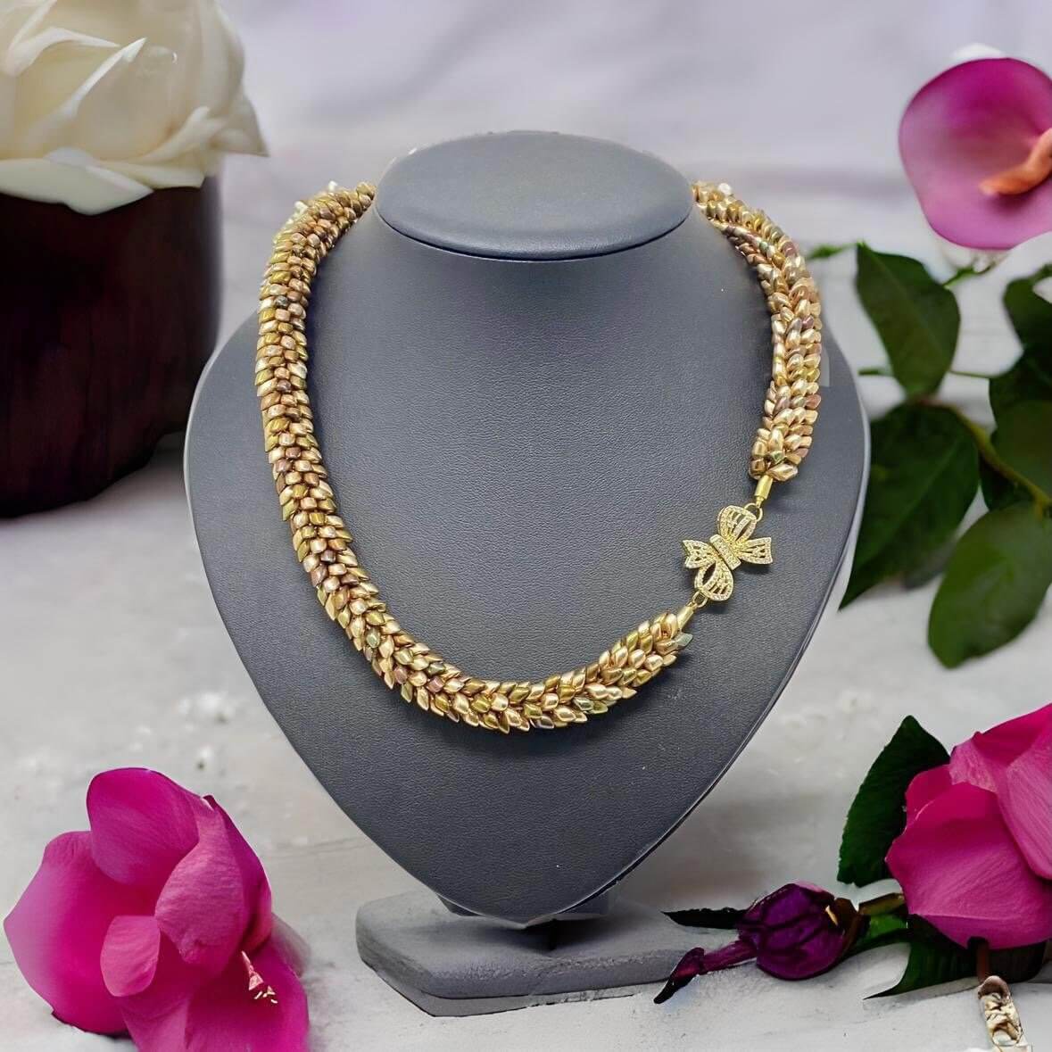 A stunning gold-toned woven necklace with a sparkling bow-shaped charm, elegantly displayed on a gray jewelry bust, surrounded by vibrant floral accents for a touch of sophistication and natural beauty.