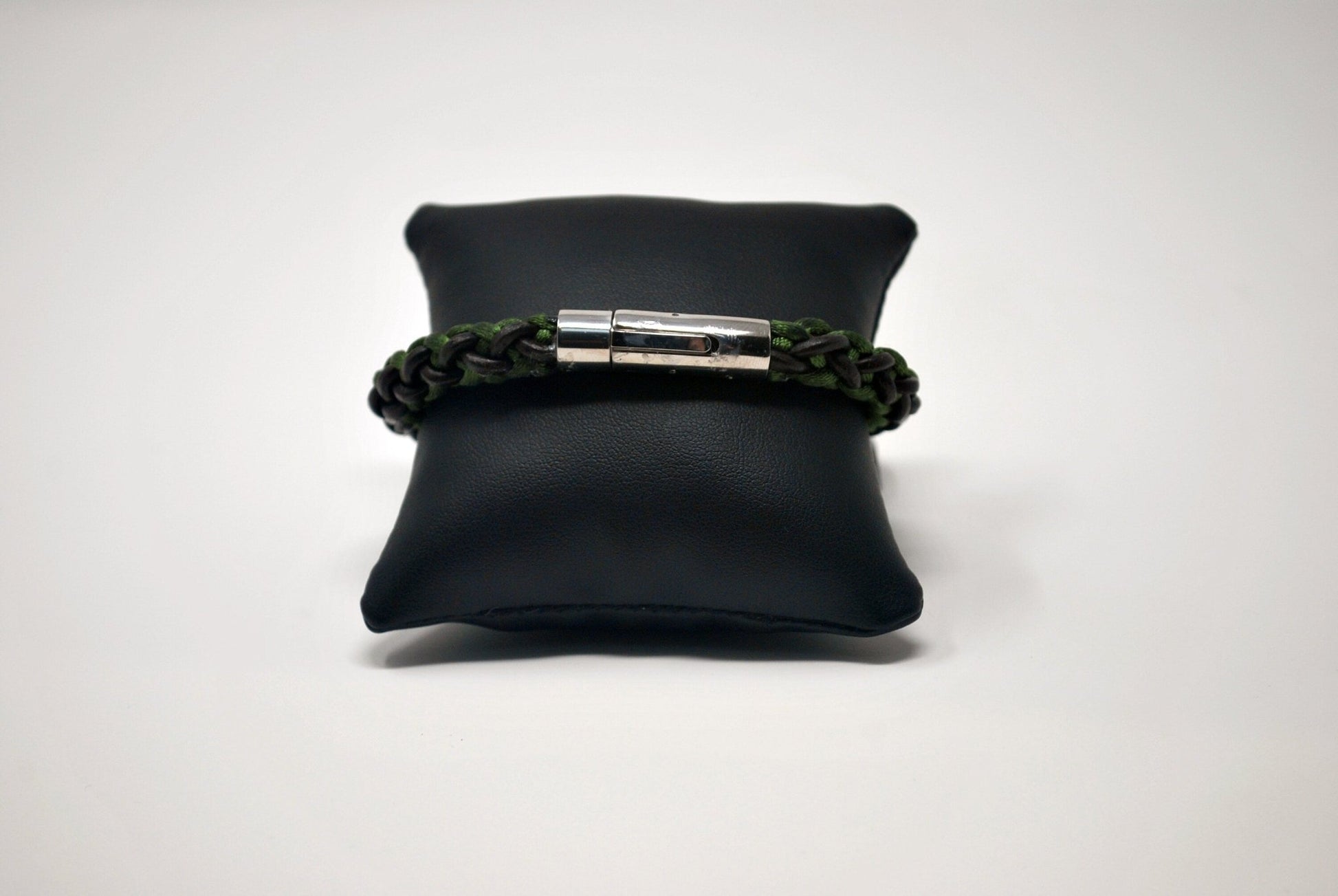 A braided bracelet with green and brown tones, featuring a stainless steel magnetic clasp, elegantly displayed on a black leather cushion against a white background.