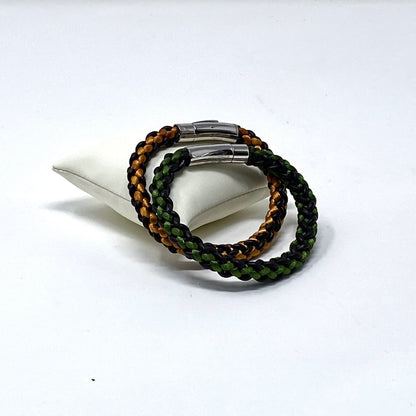 Two braided bracelets, one featuring green and brown tones and the other orange and brown, both with stainless steel magnetic clasps, elegantly arranged on an ivory leather cushion and displayed on a white background.