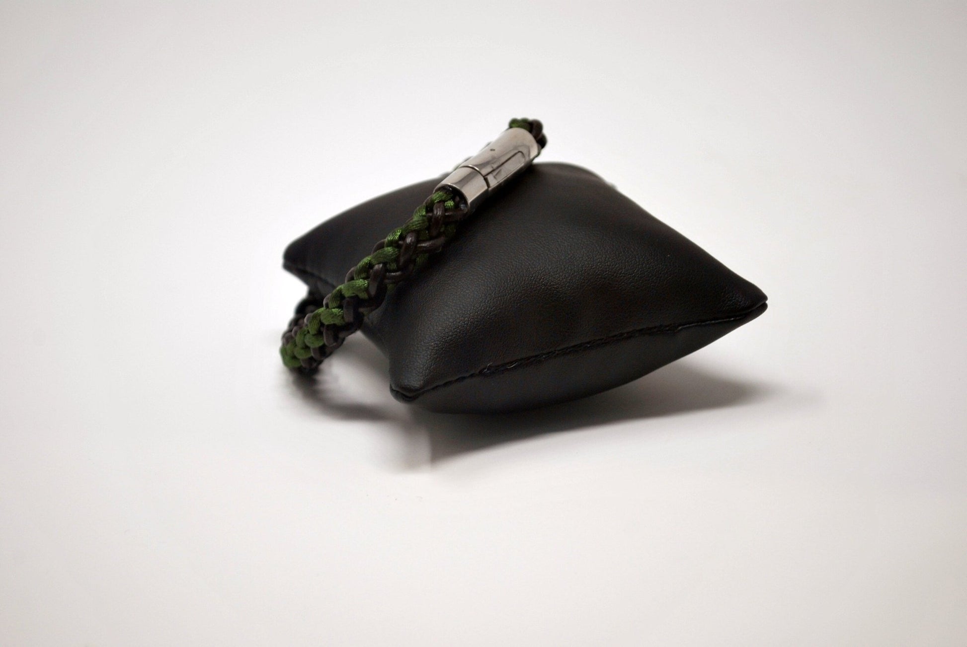 A braided bracelet with green and brown tones, featuring a stainless steel magnetic clasp, elegantly displayed on a black leather cushion against a white background.